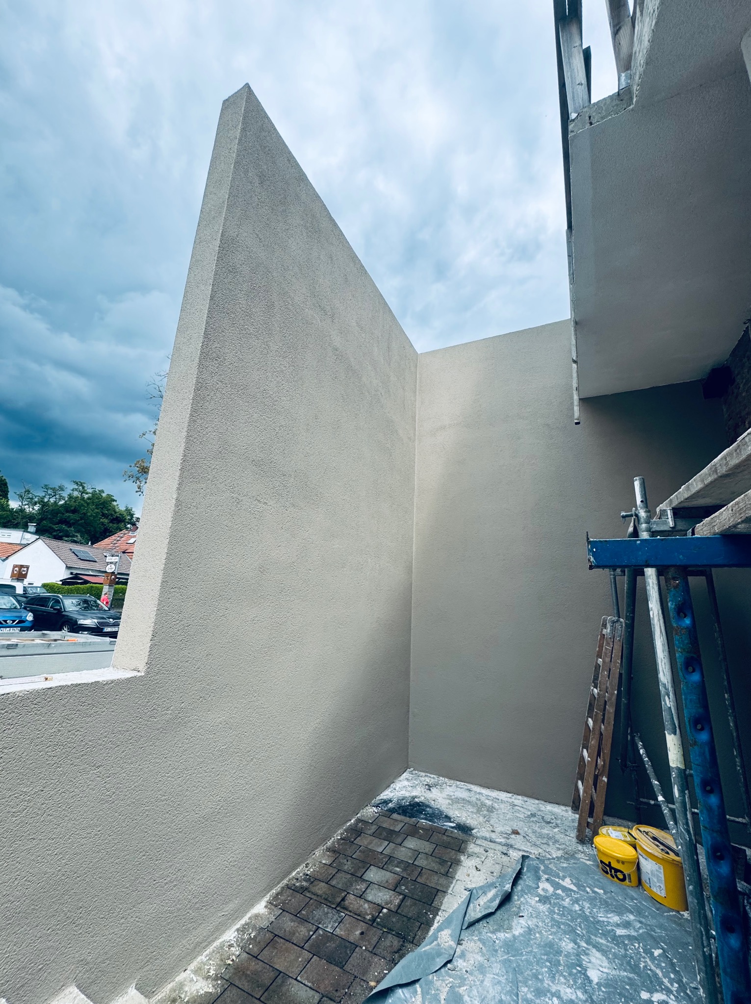 Świeżo otynkowany balkon z wykończoną elewacją w neutralnym odcieniu szarości. Widoczne rusztowania i narzędzia malarskie. Tło stanowi pochmurne niebo i zabudowa miejska.