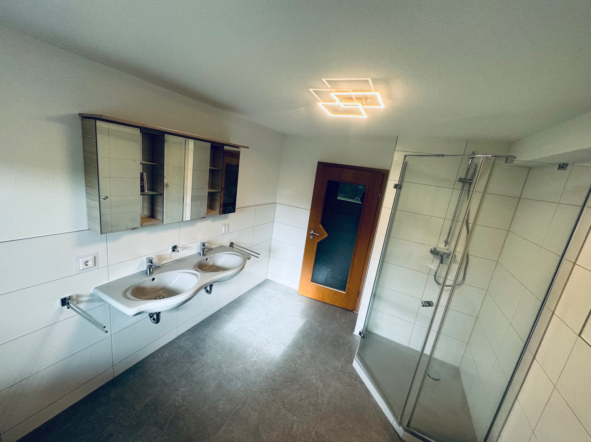 Jasna łazienka z dwoma umywalkami, kabiną prysznicową i lustrem z szafką. Uwagę przykuwa nowoczesna lampa sufitowa i drewniane drzwi.