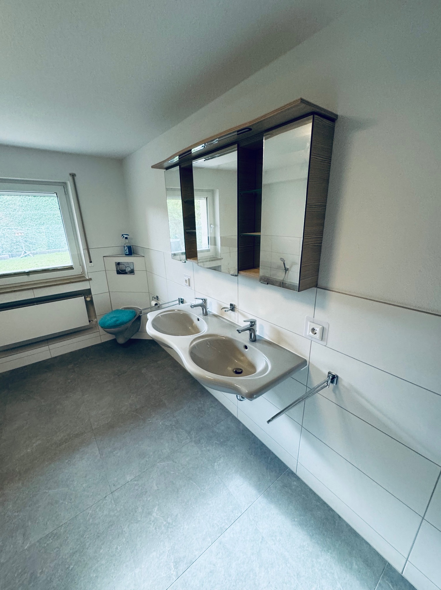 Jasna łazienka z podwójną umywalką i szafką z lustrem. Minimalistyczny design, szare płytki podłogowe i białe ściany. Widok na okno z zielenią na zewnątrz.