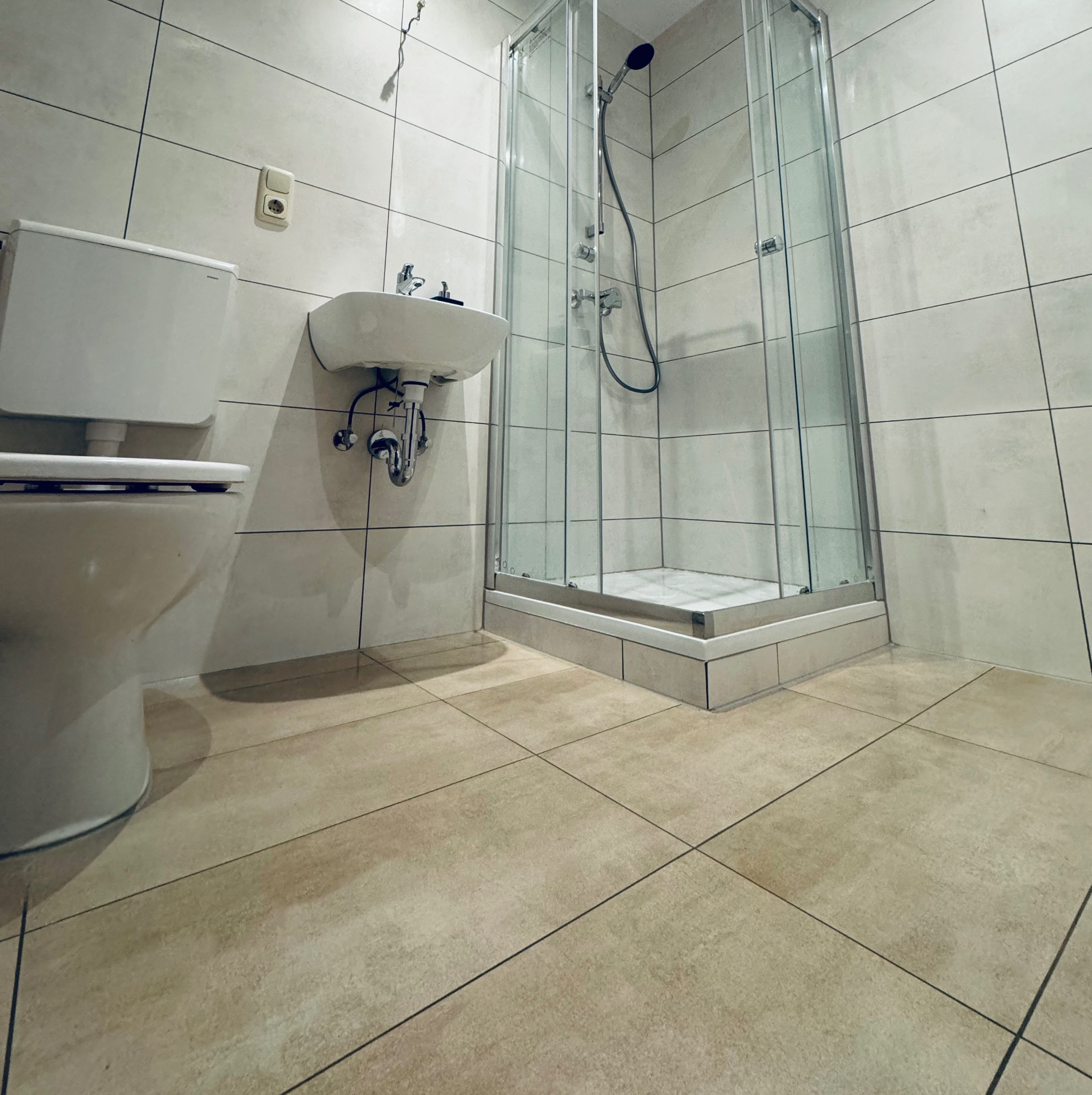 Jasna łazienka po remoncie: kabina prysznicowa, umywalka i toaleta z beżowymi płytkami podłogowymi i szarymi ściennymi. Widoczne gniazdko elektryczne.