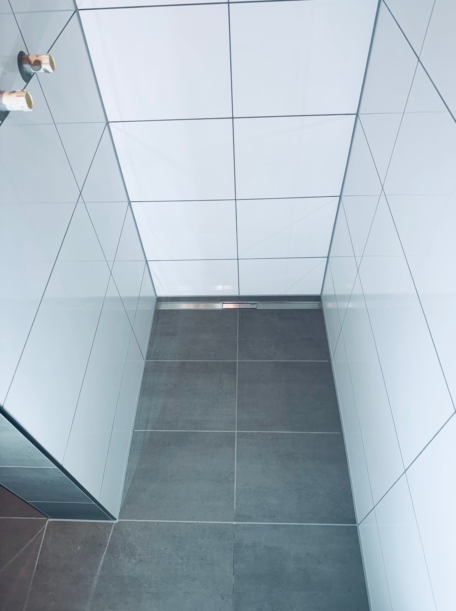 Minimalistyczna łazienka z szarymi płytkami podłogowymi i białymi ściennymi. Widoczny odpływ liniowy i fragment armatury. Ujęcie z góry, podkreślające geometrię.