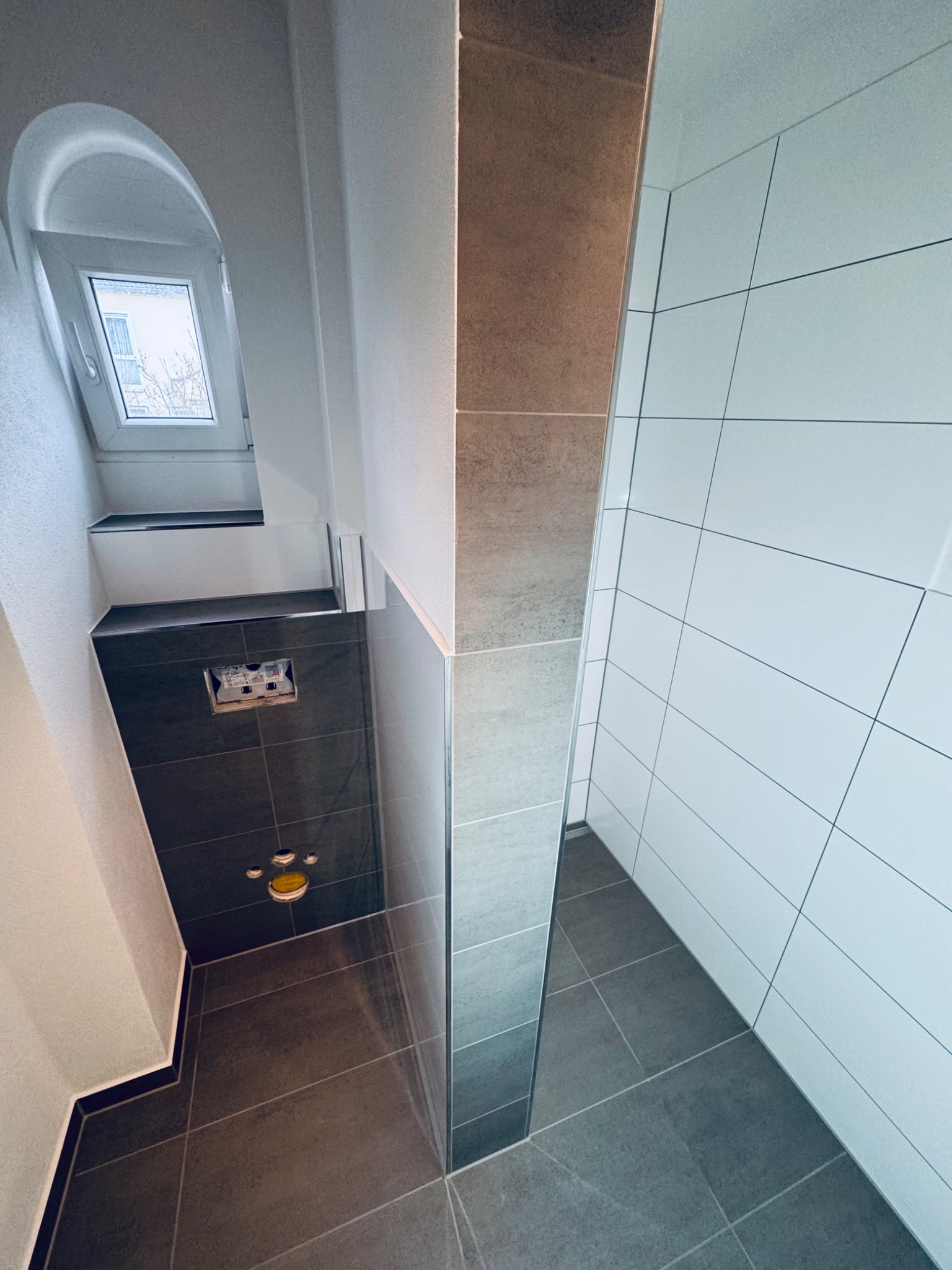 Wykończona łazienka z oknem w kształcie łuku. Ciemne i jasne płytki ceramiczne na ścianach i podłodze. Widoczne elementy instalacji sanitarnej.