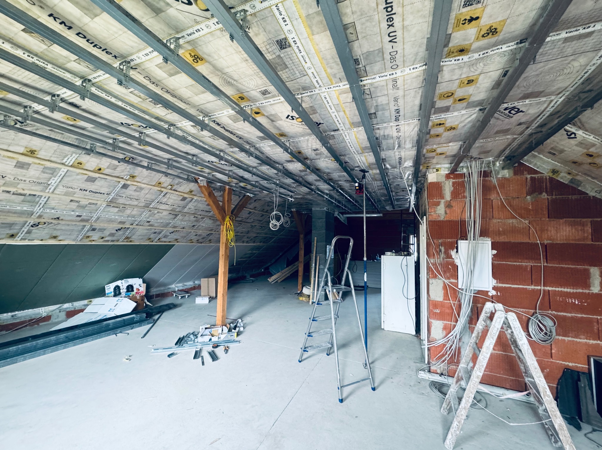 Poddasze w trakcie adaptacji: widoczne krokwie, izolacja, metalowe profile sufitowe i instalacje elektryczne przy ceglanej ścianie. Dwie drabiny i materiały budowlane na podłodze.