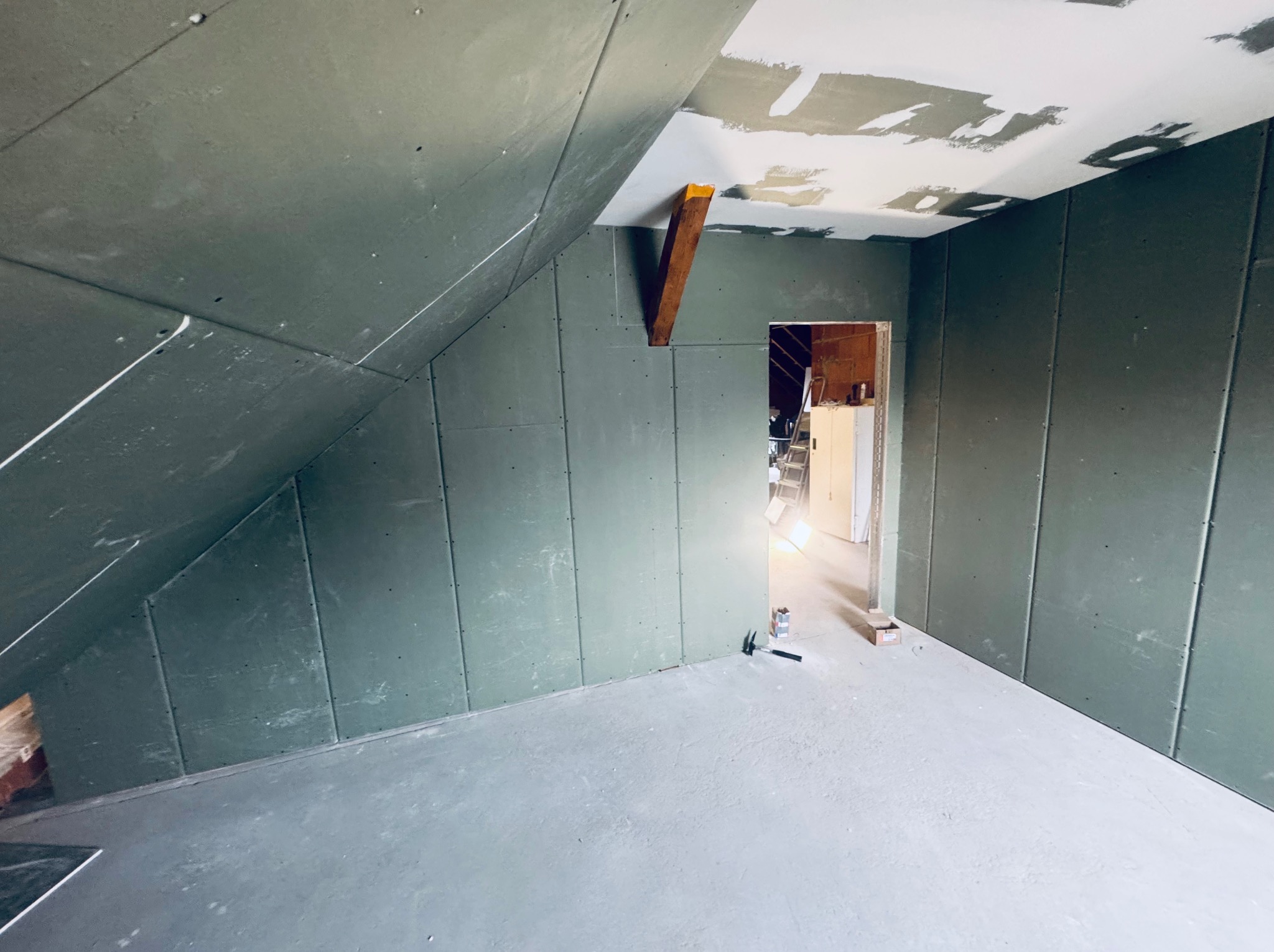 Poddasze w trakcie adaptacji: ściany i skosy obłożone płytami gipsowo-kartonowymi, widoczne łączenia płyt, sufit częściowo zagruntowany. W tle otwór drzwiowy.