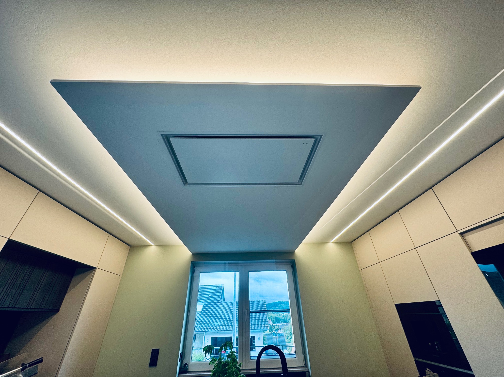 Nowoczesna kuchnia z oknem: sufit z wbudowanym okapem i oświetleniem LED. Jasne szafki kuchenne i widok na zewnątrz. Minimalistyczny design.