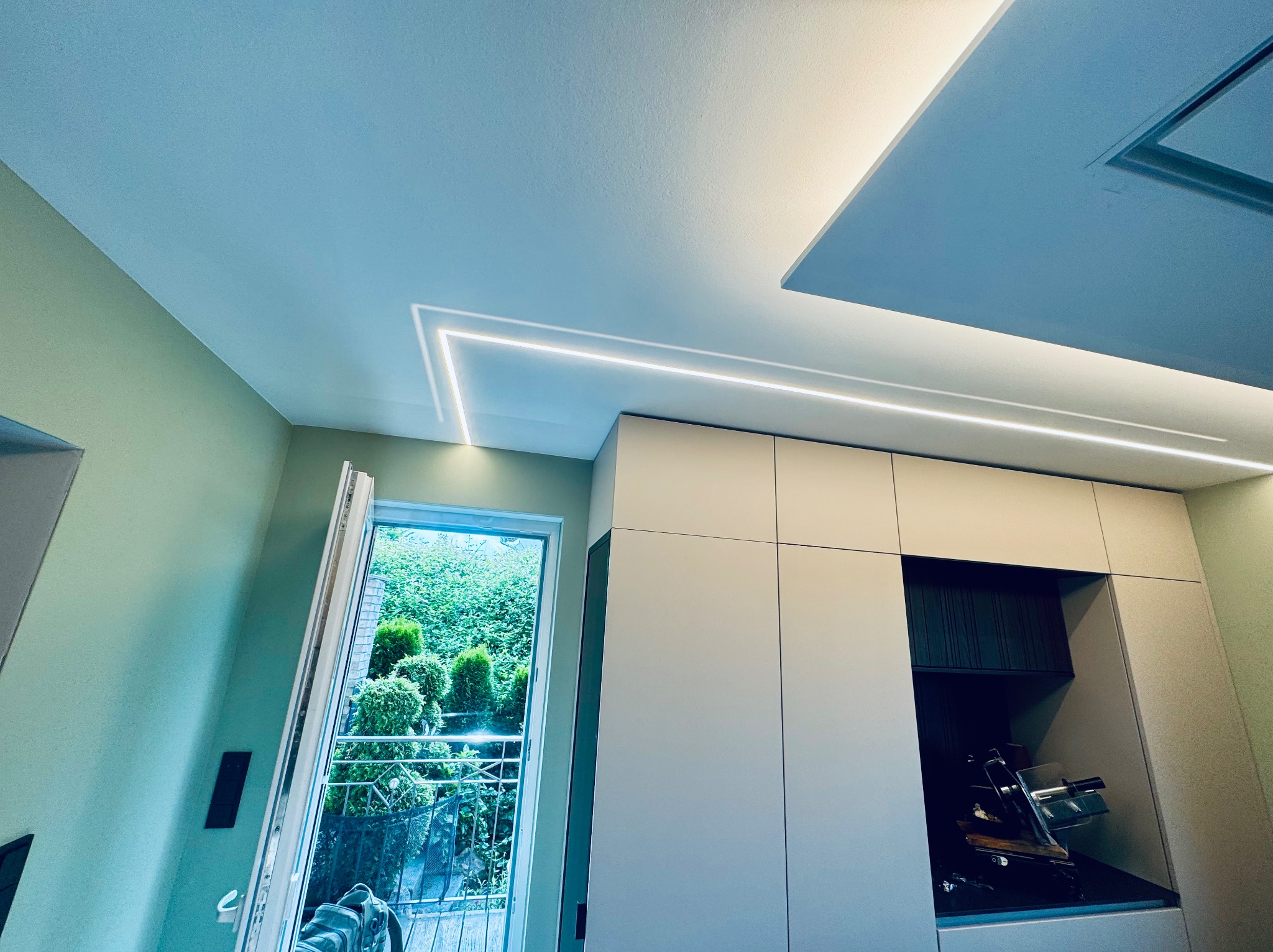 Nowoczesne oświetlenie LED w suficie, białe szafki kuchenne i otwarte drzwi balkonowe z widokiem na zieleń. Minimalistyczny design wnętrza z akcentem na światło.