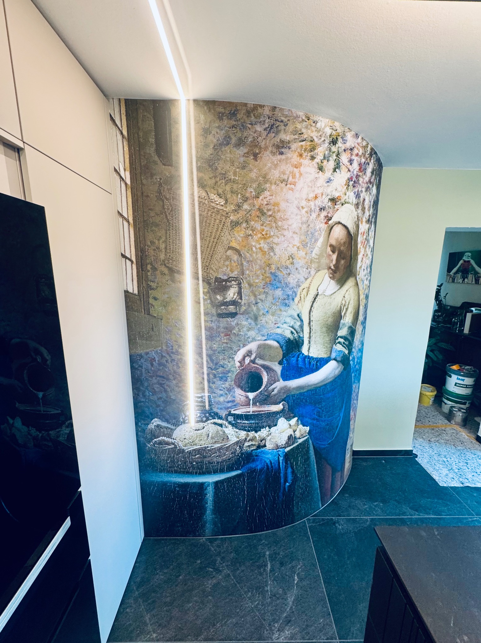 Ściana wykończona fototapetą z motywem obrazu Vermeera 'Mleczarka', wkomponowana w nowoczesną zabudowę kuchenną z oświetleniem LED. Ciemna podłoga z płytek.