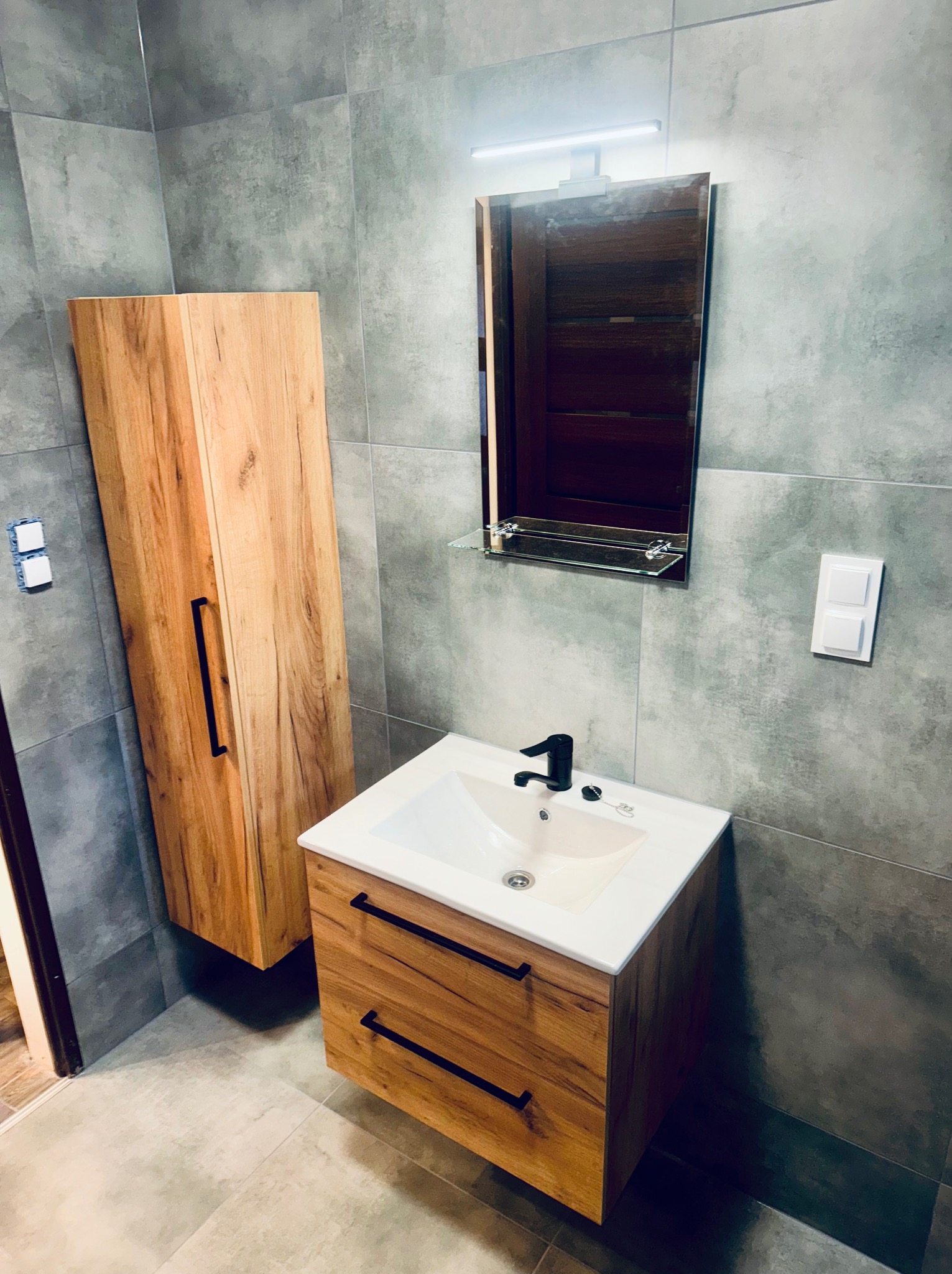 Nowoczesna łazienka z drewnianą szafką pod umywalką i wysoką szafką wiszącą. Szare płytki na ścianach i podłodze. Czarna armatura i minimalistyczne oświetlenie.
