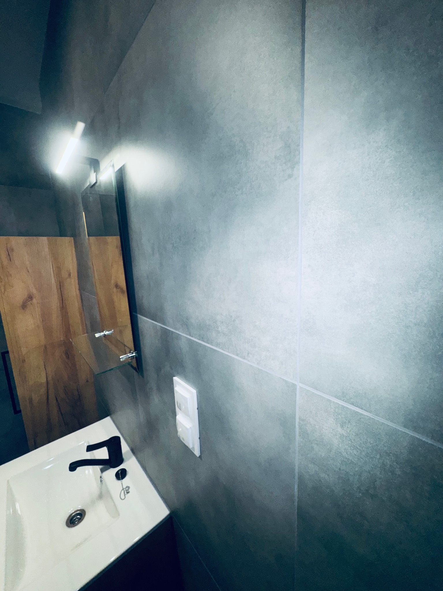 Nowoczesna łazienka w Bielawie: szare płytki, biała umywalka z czarną armaturą, lustro z oświetleniem LED i drewniane elementy wykończenia.