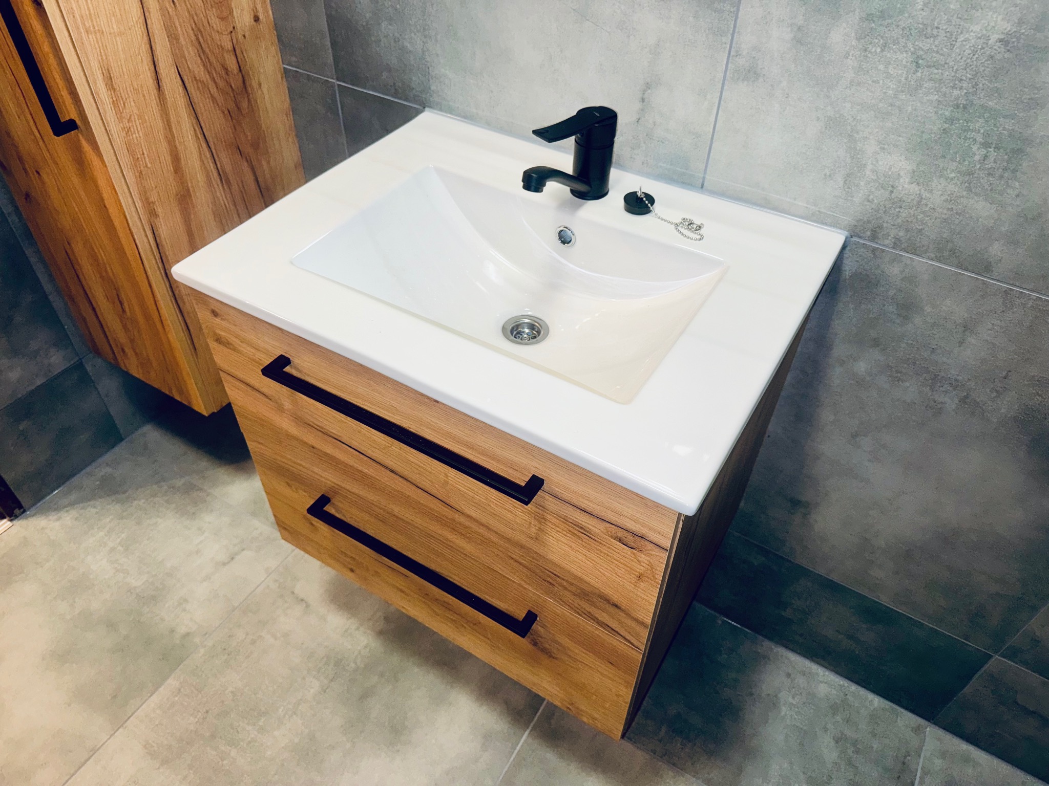 Nowoczesna szafka pod umywalkę z drewnianymi frontami i czarnymi uchwytami, biała umywalka i czarna bateria. Szare płytki na ścianie i podłodze.