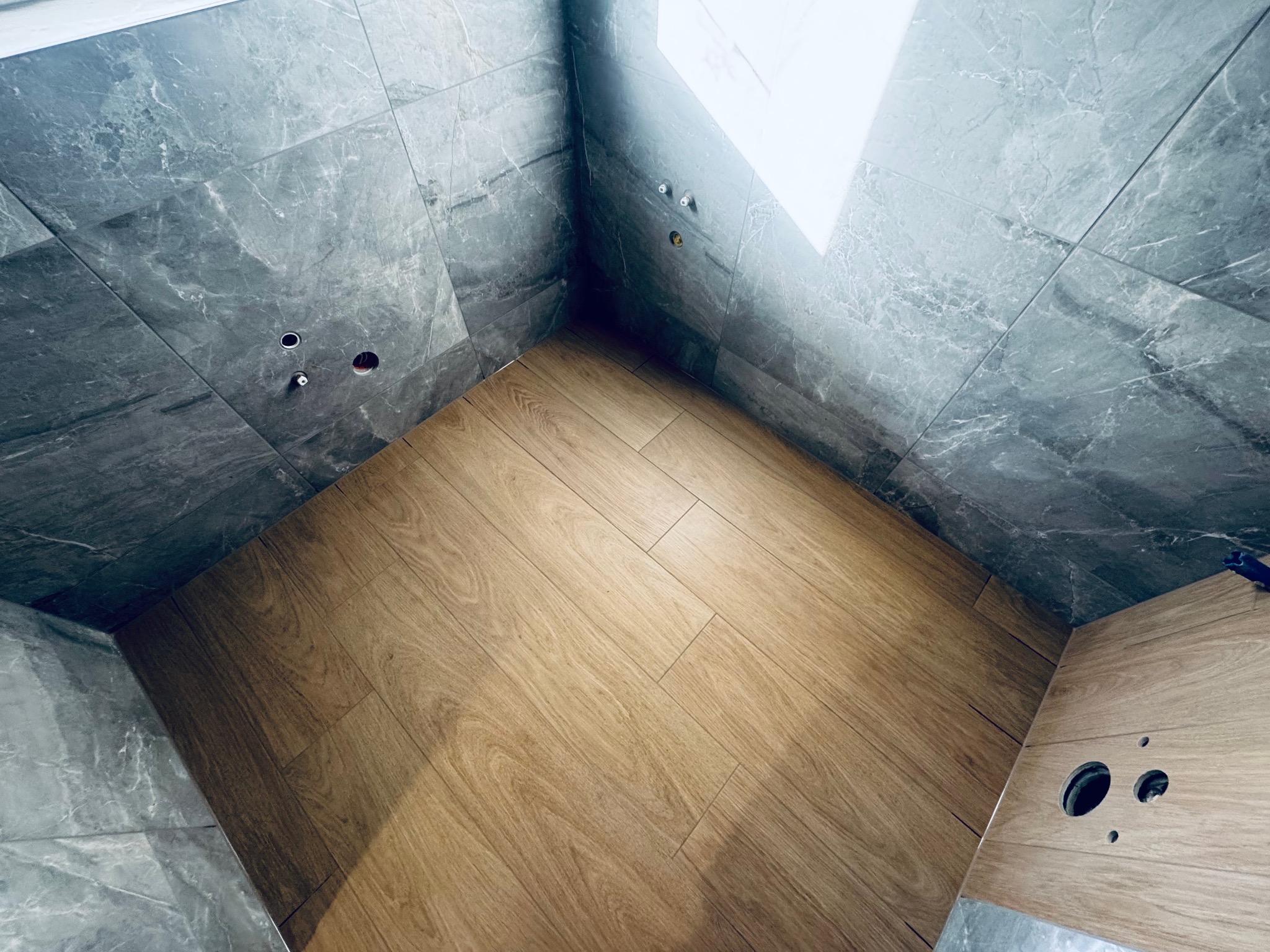 Wykończona łazienka z szarymi płytkami imitującymi marmur na ścianach i drewnopodobną podłogą. Widoczne otwory na armaturę i odpływ.