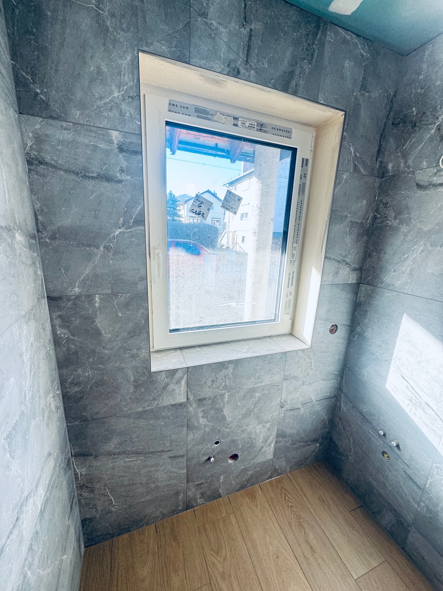 Łazienka w trakcie wykańczania: szare płytki imitujące marmur na ścianach, drewnopodobne panele na podłodze, białe okno z widokiem na domy. Widoczne otwory na armaturę.