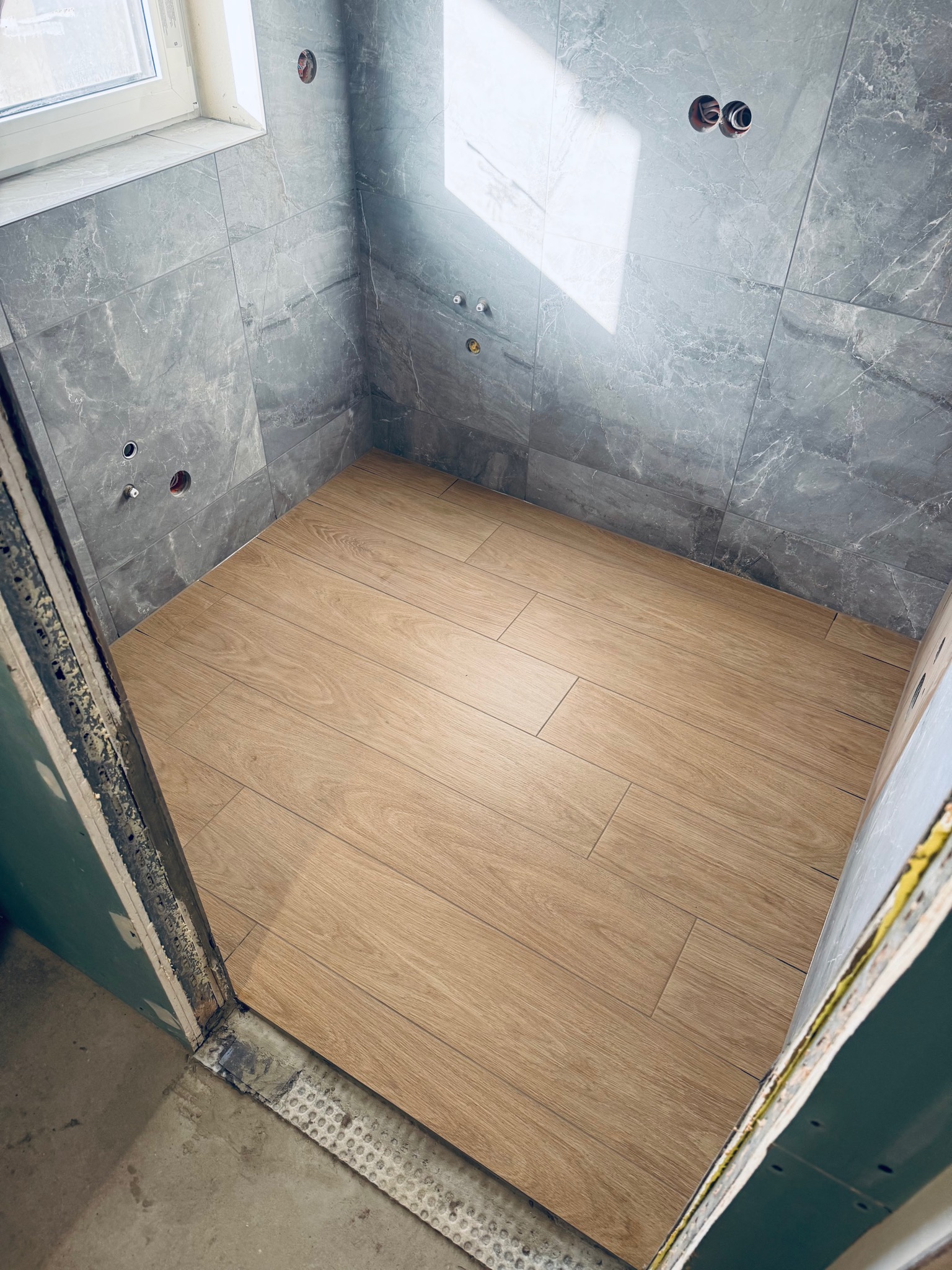 Wykończona kabina prysznicowa z szarymi płytkami imitującymi marmur na ścianach i drewnopodobną podłogą. Widoczne otwory na armaturę i surowe wykończenie wejścia.