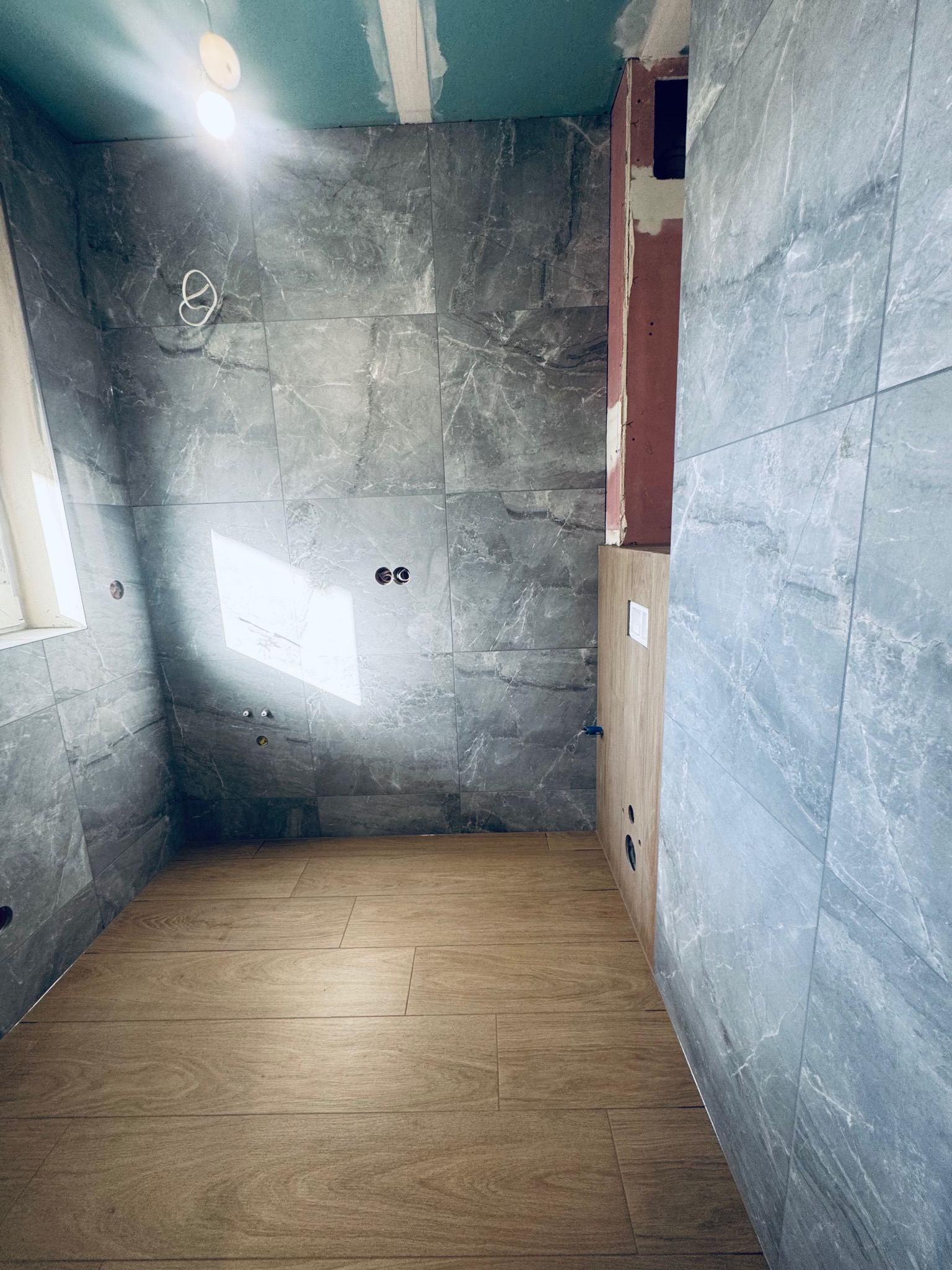 Wnętrze łazienki w trakcie remontu: szare płytki imitujące marmur na ścianach, drewnopodobna podłoga, widoczne instalacje elektryczne i hydrauliczne, sufit z płyt gipsowych.