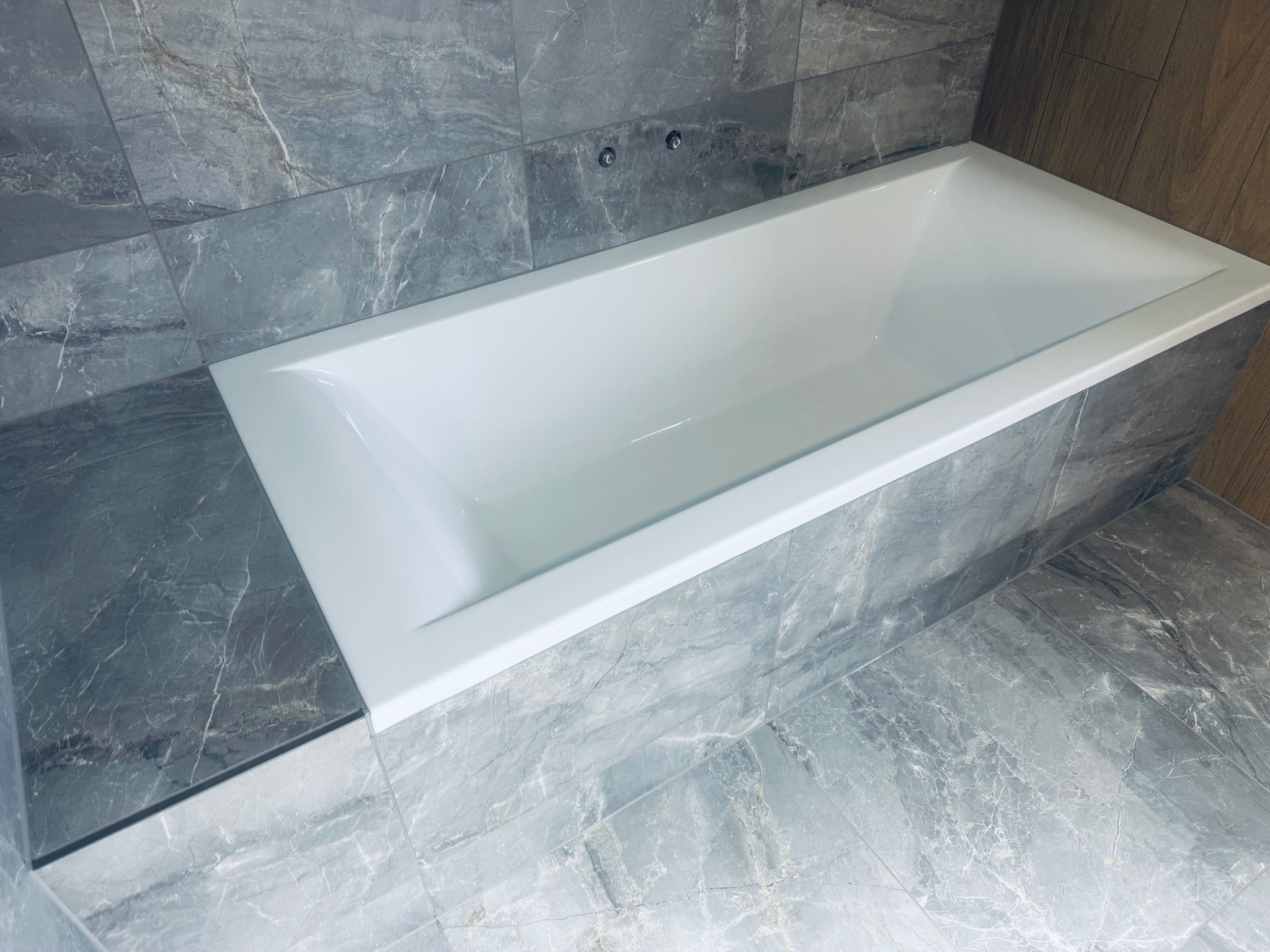 Nowoczesna, wykończona łazienka z białą wanną i szarymi płytkami imitującymi marmur na ścianach i podłodze. Widoczne chromowane elementy armatury.