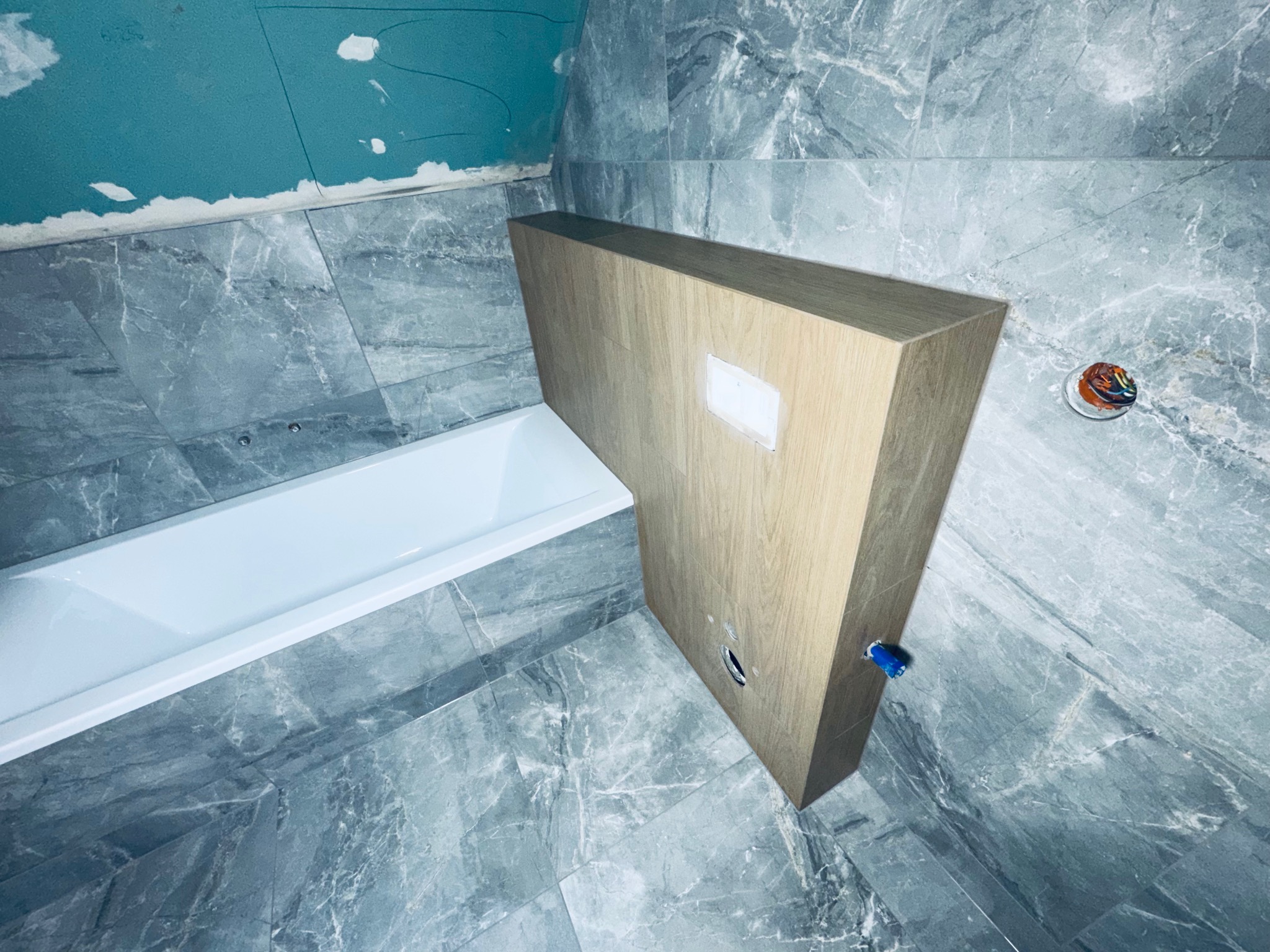 Łazienka w trakcie wykończenia: szare płytki imitujące marmur, zabudowa wanny z drewnopodobnych paneli, widoczne elementy instalacji. Surowy stan, niebieska ściana.