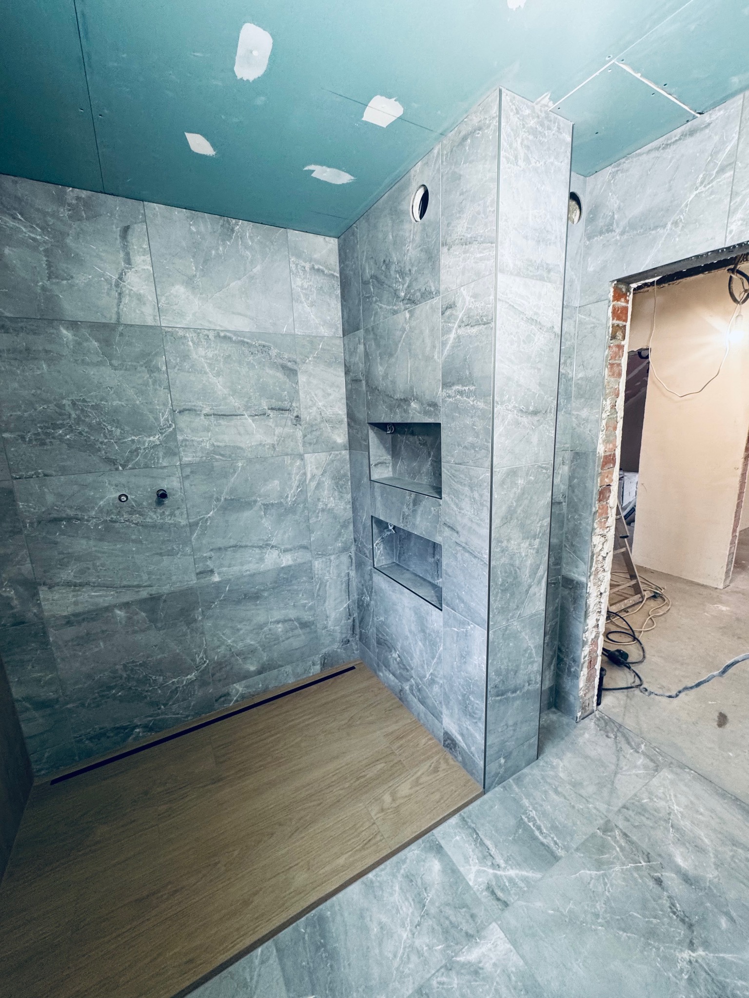 Łazienka w trakcie wykończenia: szare płytki imitujące marmur na ścianach i podłodze, drewnopodobny brodzik, wnęki w ścianie. Widoczne niedokończone prace.