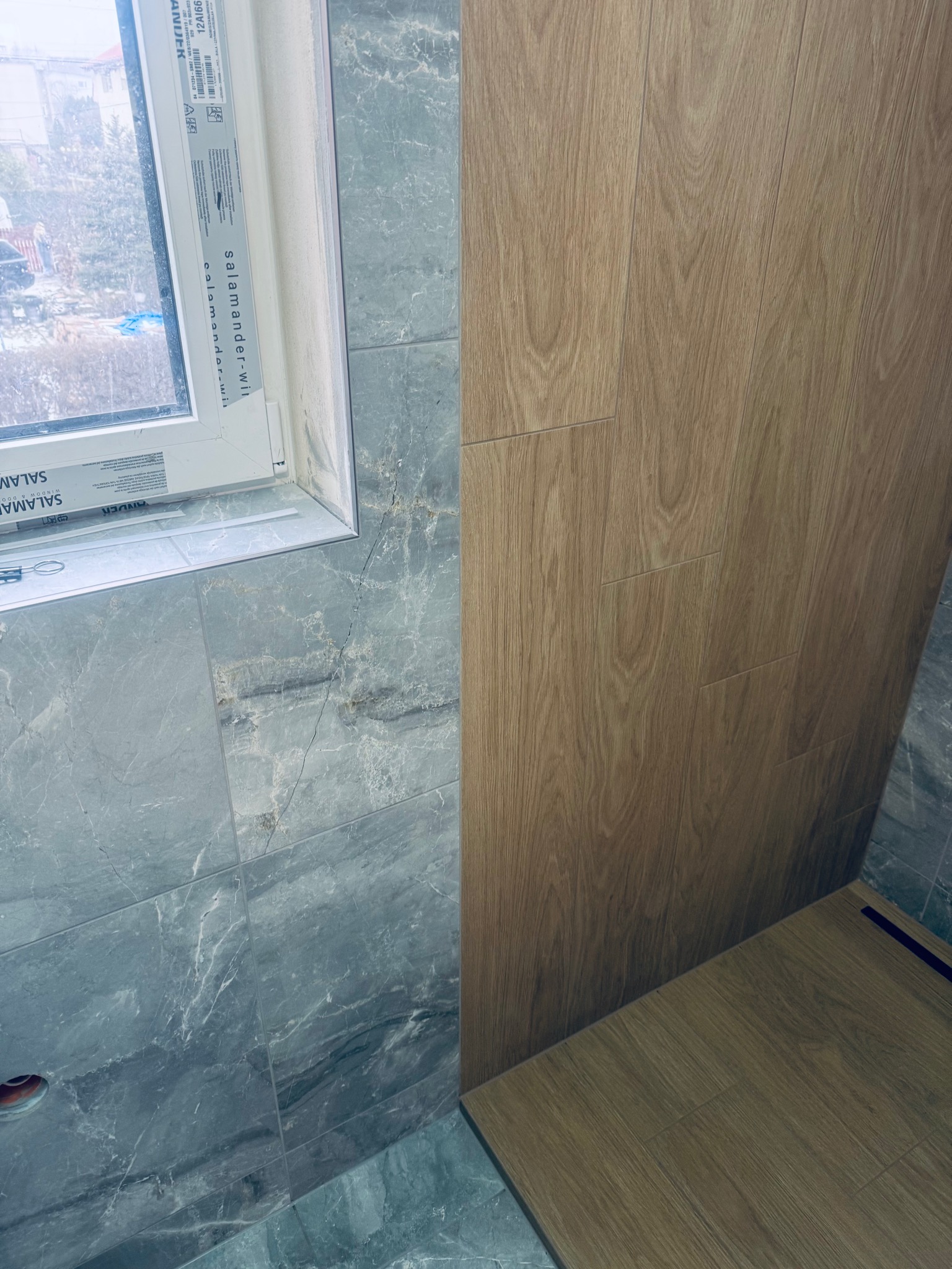 Wykończona łazienka: szare płytki imitujące marmur na ścianach i podłodze, drewnopodobne panele na ścianie, białe okno z widokiem na zewnątrz.