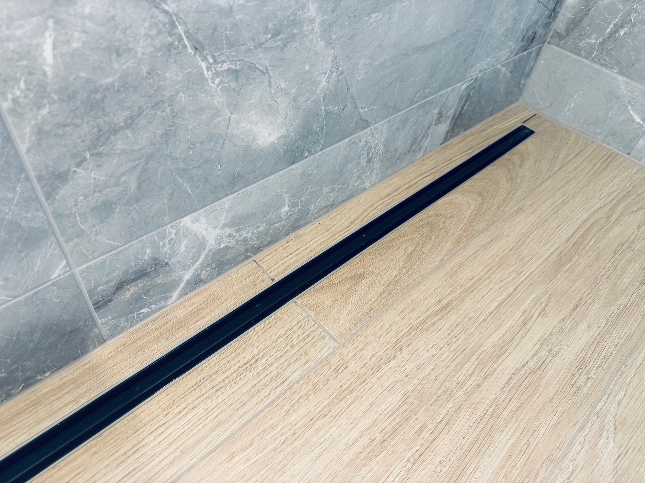 Łazienka po remoncie: jasne płytki imitujące drewno na podłodze, szare marmurowe płytki na ścianie, czarny odpływ liniowy. Minimalistyczny design.