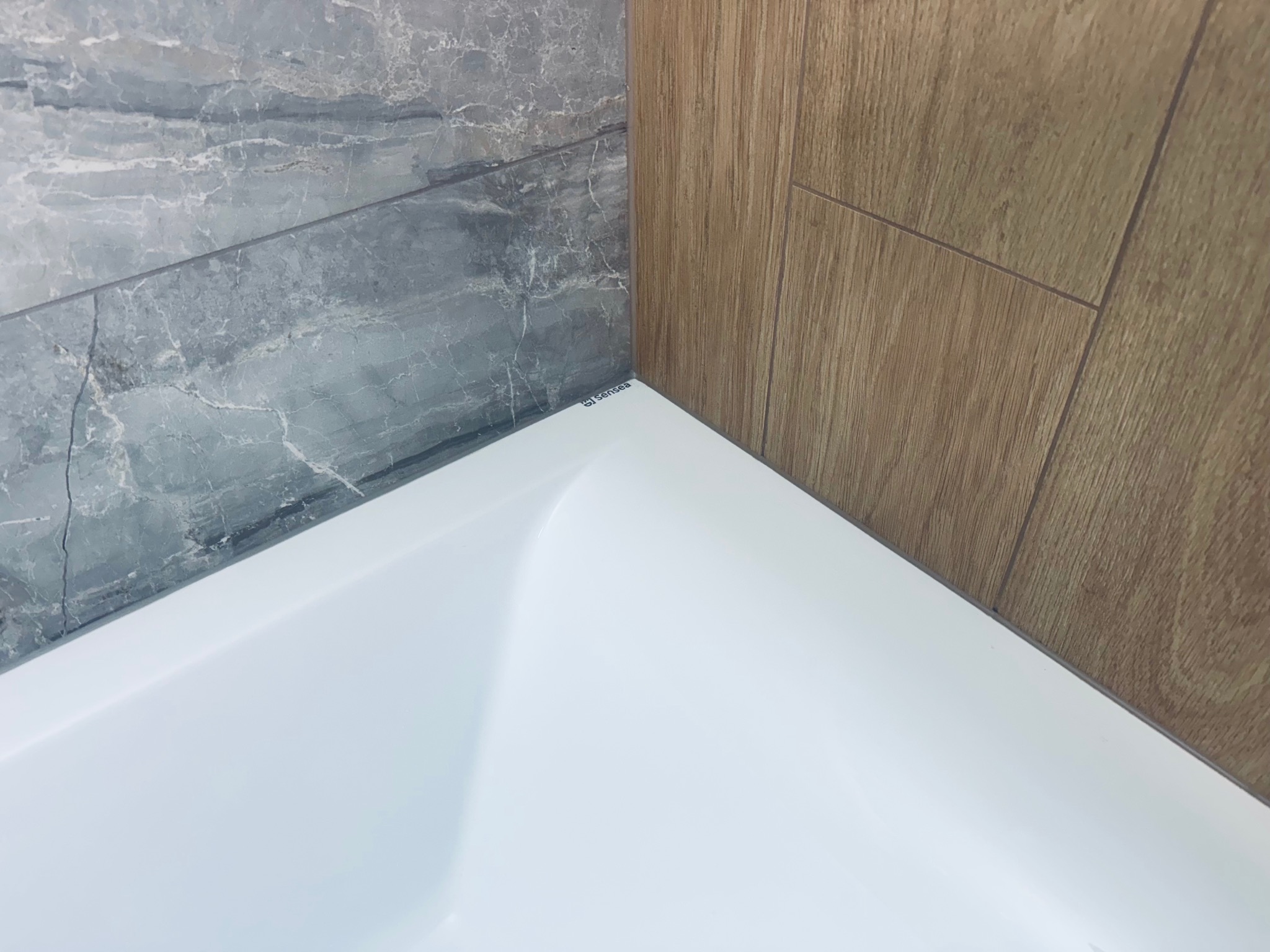 Narożnik łazienki: biała wanna, marmurowe płytki ścienne i drewnopodobne panele. Widoczny fragment wanny z logo producenta.