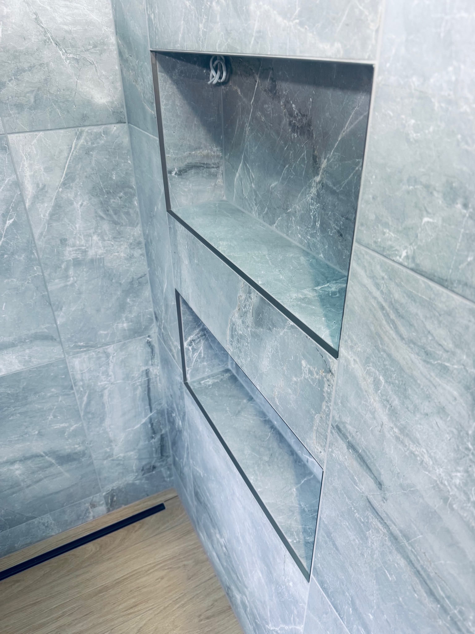 Narożna półka w łazience wyłożonej marmurowymi płytkami w odcieniach szarości. Widoczny fragment drewnianej podłogi i czarna listwa przypodłogowa.