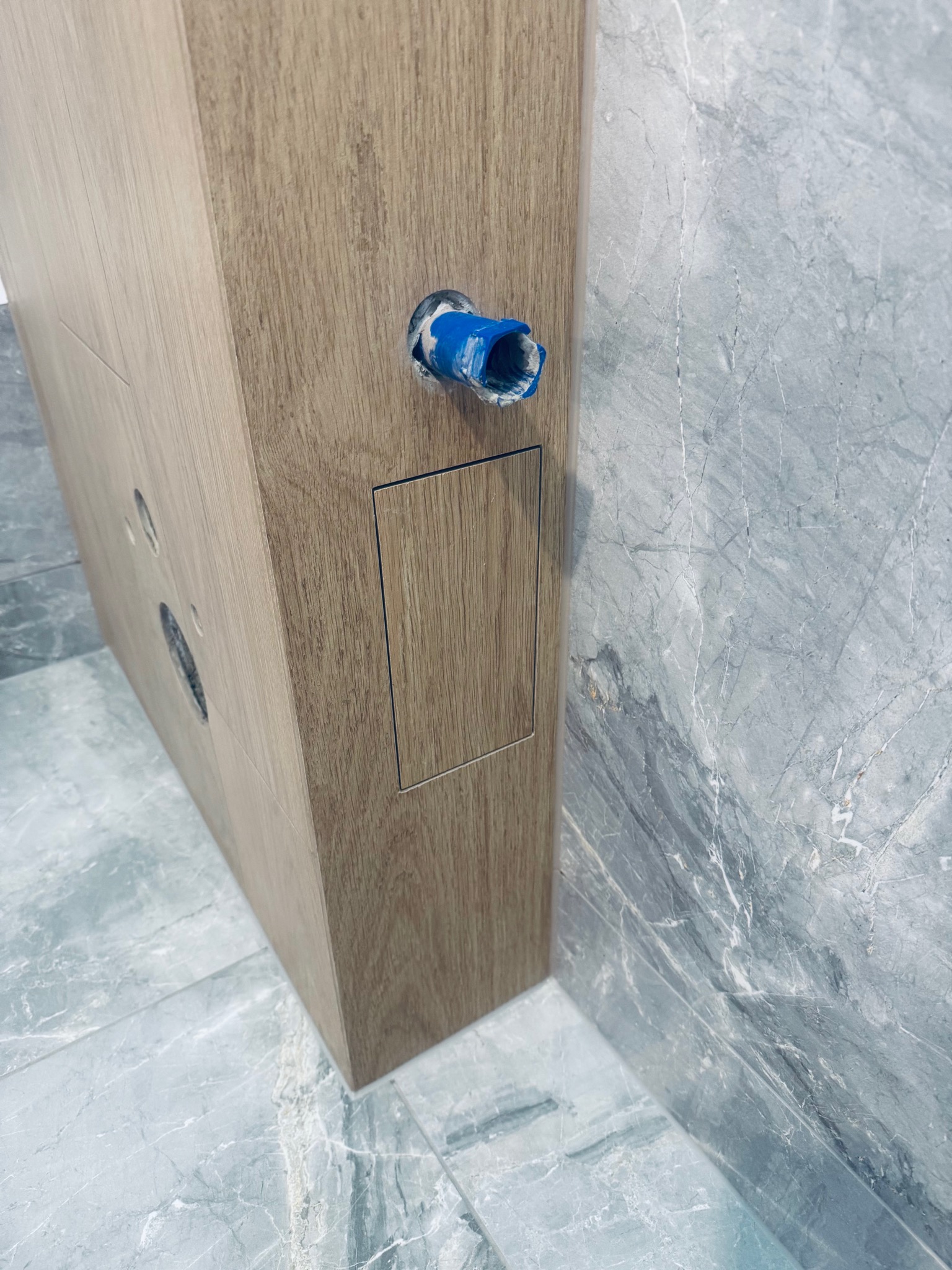 Drewniana obudowa stelaża WC z otworem na rurę, na tle marmurowej ściany i podłogi. Widoczne detale wykończenia i instalacji w łazience.