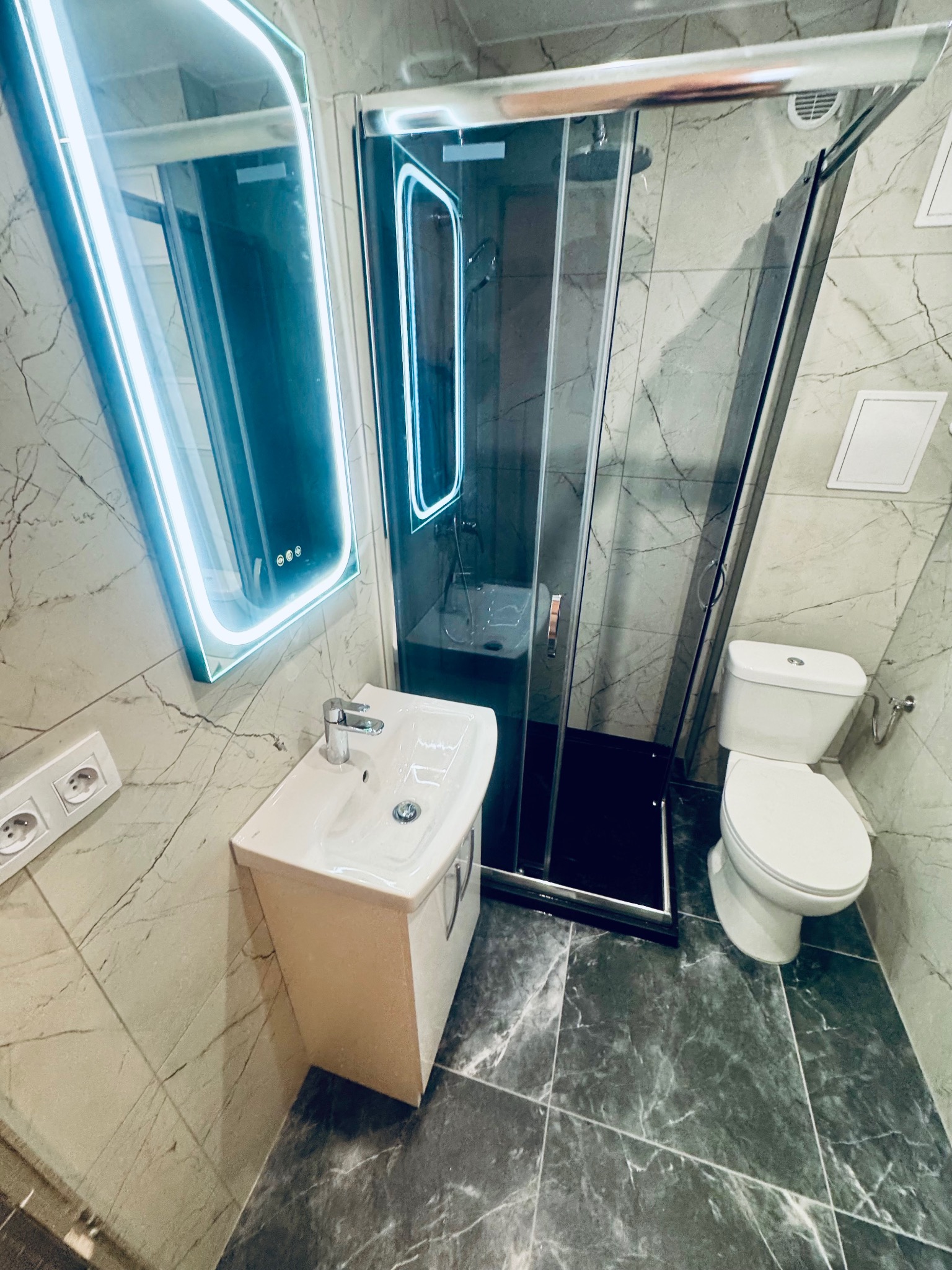 Wykończona łazienka z kabiną prysznicową, umywalką z szafką i toaletą. Ściany i podłoga wyłożone płytkami imitującymi marmur. Lustro z podświetleniem LED.