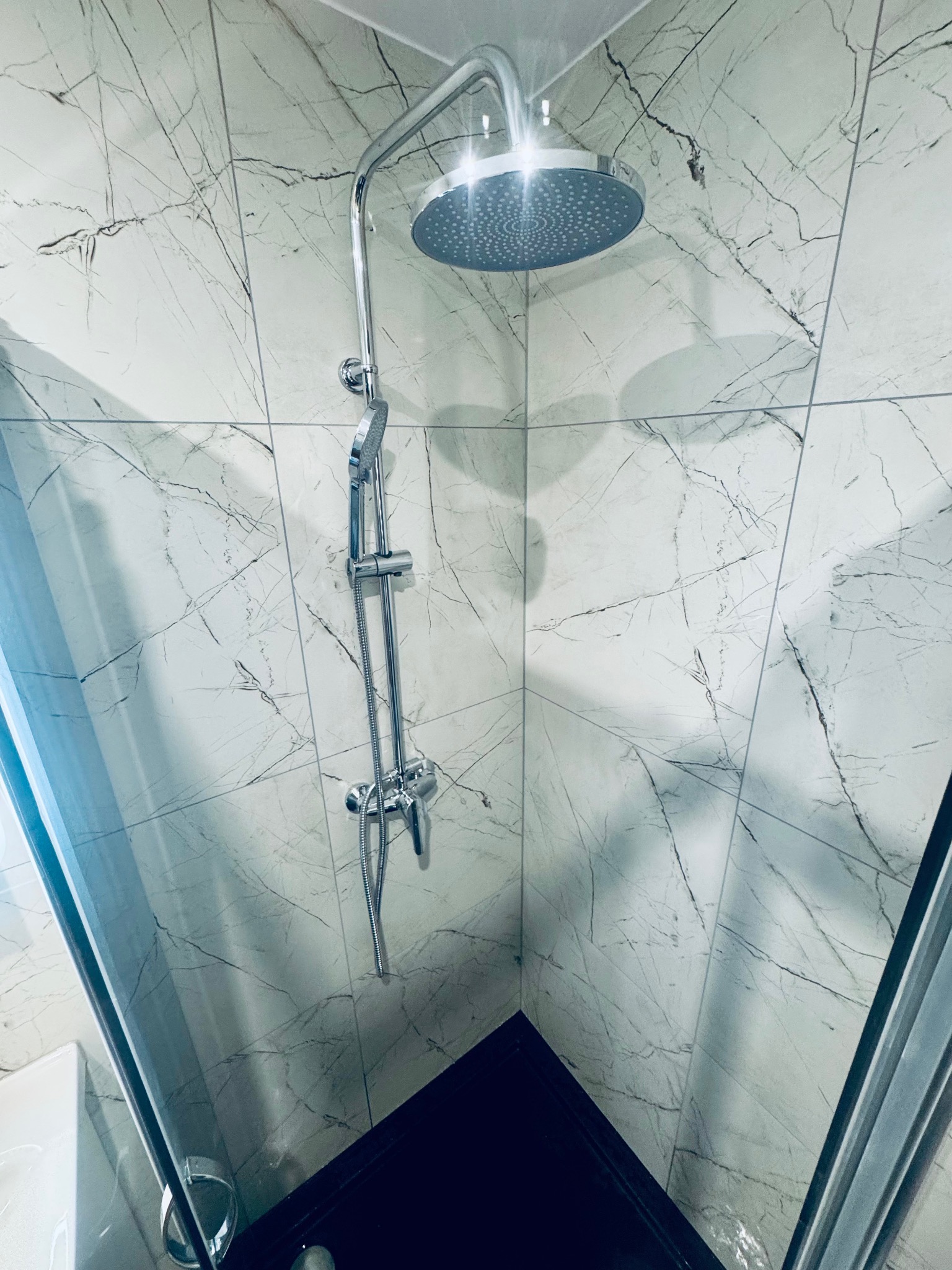 Nowoczesna kabina prysznicowa z deszczownicą i marmurowymi płytkami. Chromowana armatura błyszczy w świetle. Widoczny fragment czarnego brodzika i szklane drzwi.