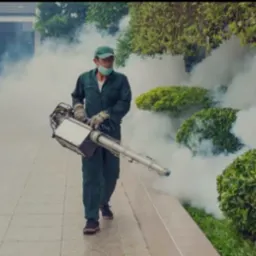 Pracownik w kombinezonie ochronnym rozpyla środek zamgławiający na krzewy przy budynku, używając specjalistycznego sprzętu. Biała mgła unosi się w powietrzu.