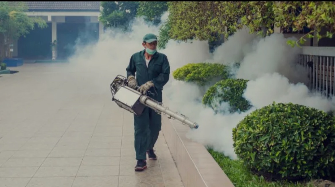 Pracownik w kombinezonie ochronnym rozpyla środek zamgławiający na krzewy przy budynku, używając specjalistycznego sprzętu. Biała mgła unosi się w powietrzu.