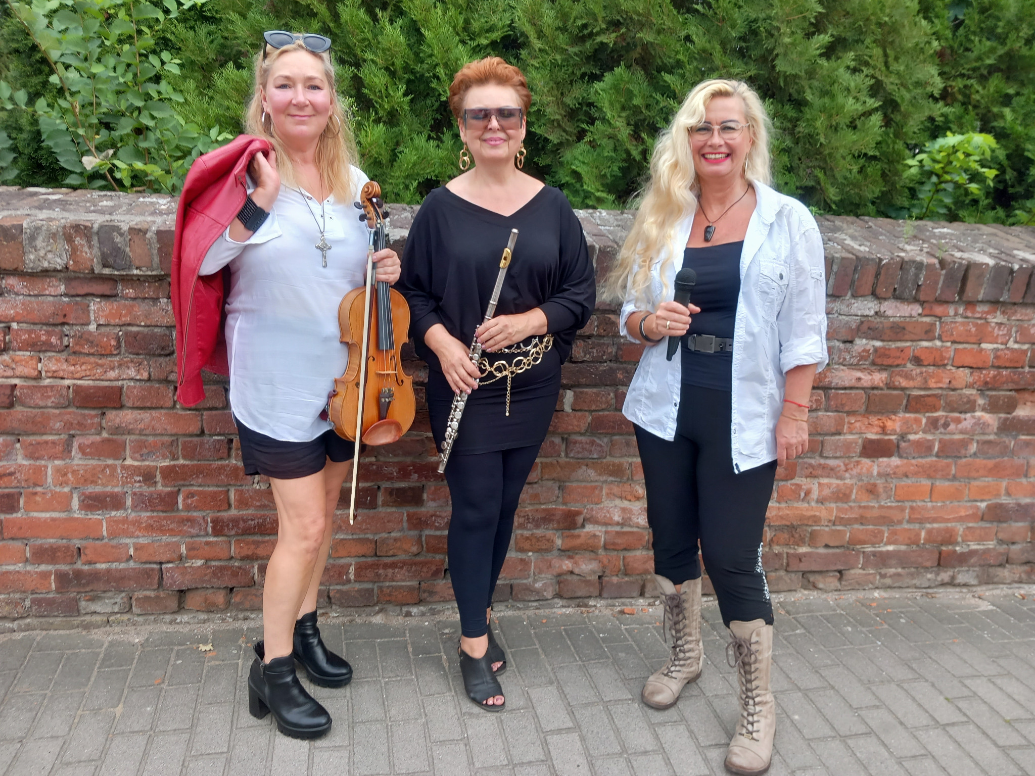 Trzy kobiety, muzyczne trio, prezentują instrumenty: skrzypce, flet i mikrofon. Ceglana ściana i zieleń w tle. Różne style ubioru, od swobodnego do eleganckiego.