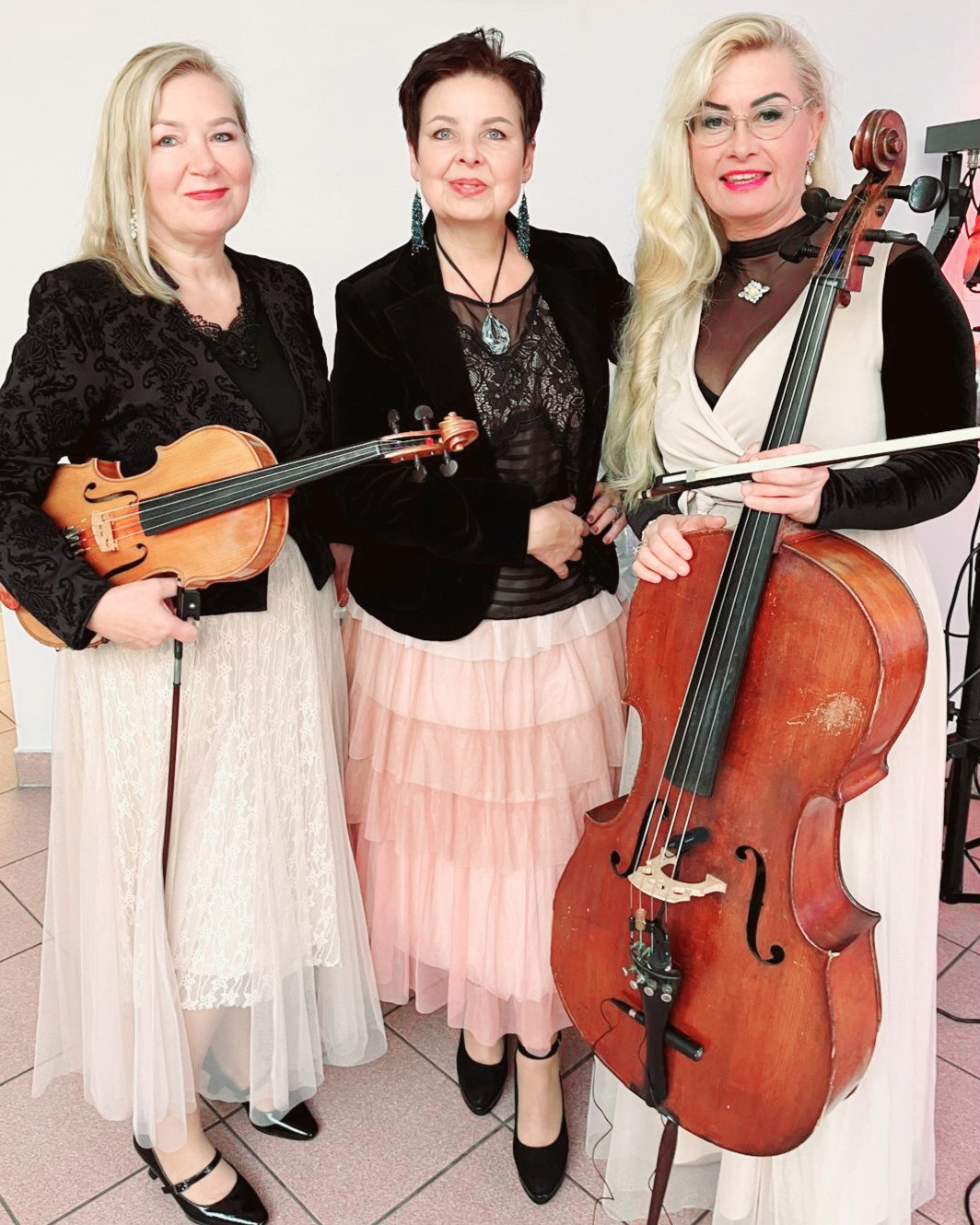 Elegancki damski zespół smyczkowy w stylizowanych sukniach. Dwie skrzypaczki i wiolonczelistka prezentują instrumenty. Scena w jasnych barwach.