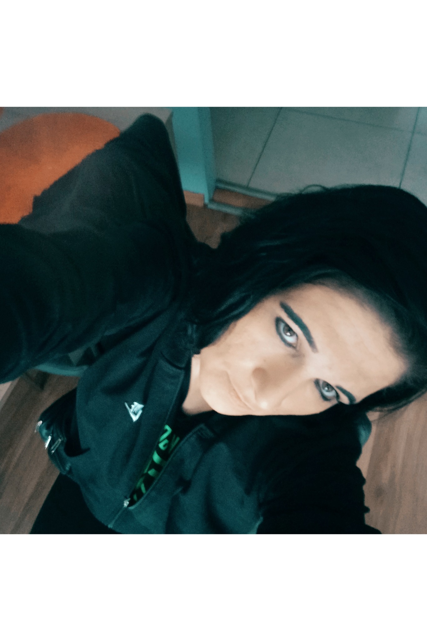 Kobieta z ciemnymi włosami w czarnej bluzie, patrzy w dół, widok z góry. Fragment podłogi i krzesła w tle. Wyraźny makijaż oczu.