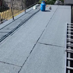 Wykończony dach pokryty papą termozgrzewalną z widocznym zbiornikiem gazu i palnikiem. Metalowa balustrada i drabina. Widok na zieleń i zabudowę.