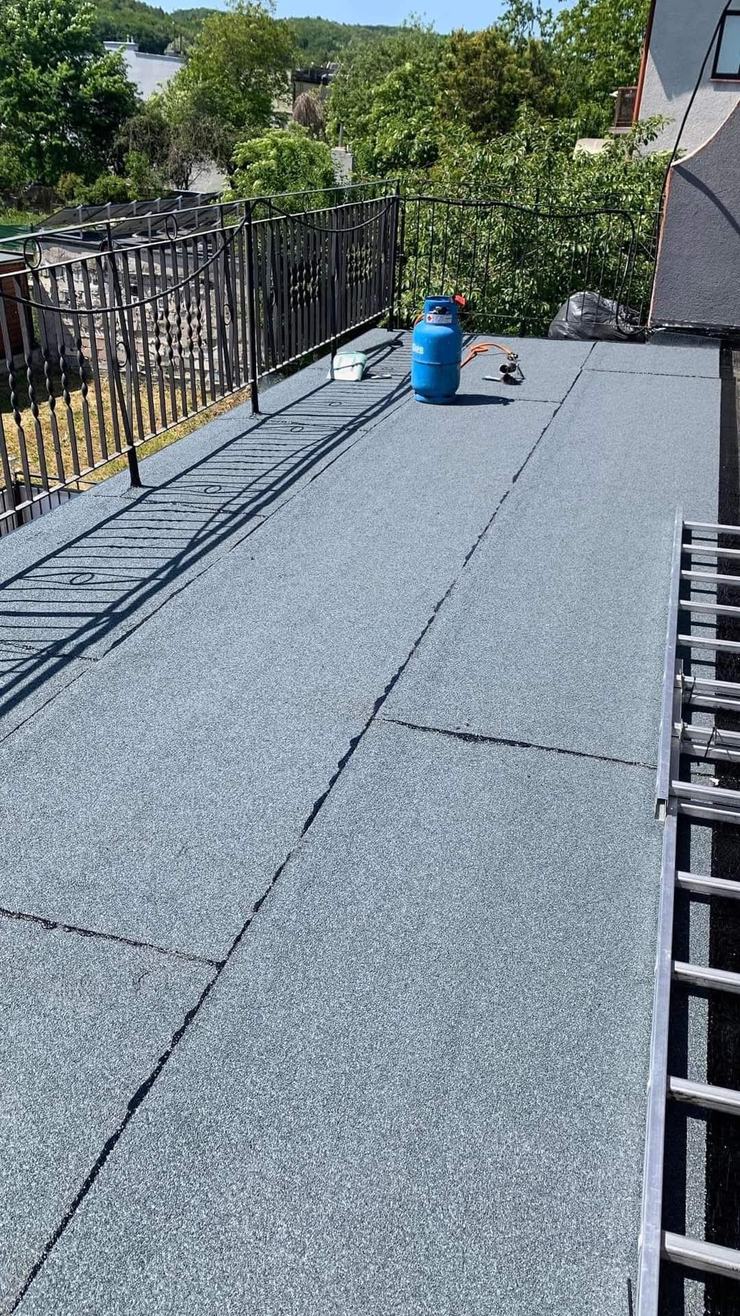 Wykończony dach pokryty papą termozgrzewalną z widocznym zbiornikiem gazu i palnikiem. Metalowa balustrada i drabina. Widok na zieleń i zabudowę.