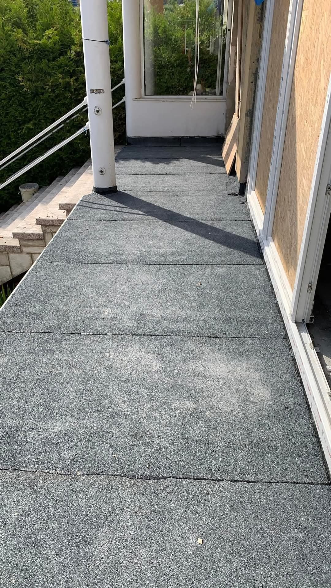 Wykończony płaski dach z szarej papy termozgrzewalnej, widok z góry, częściowo zadaszony, z kolumną i oknami, w tle zieleń.