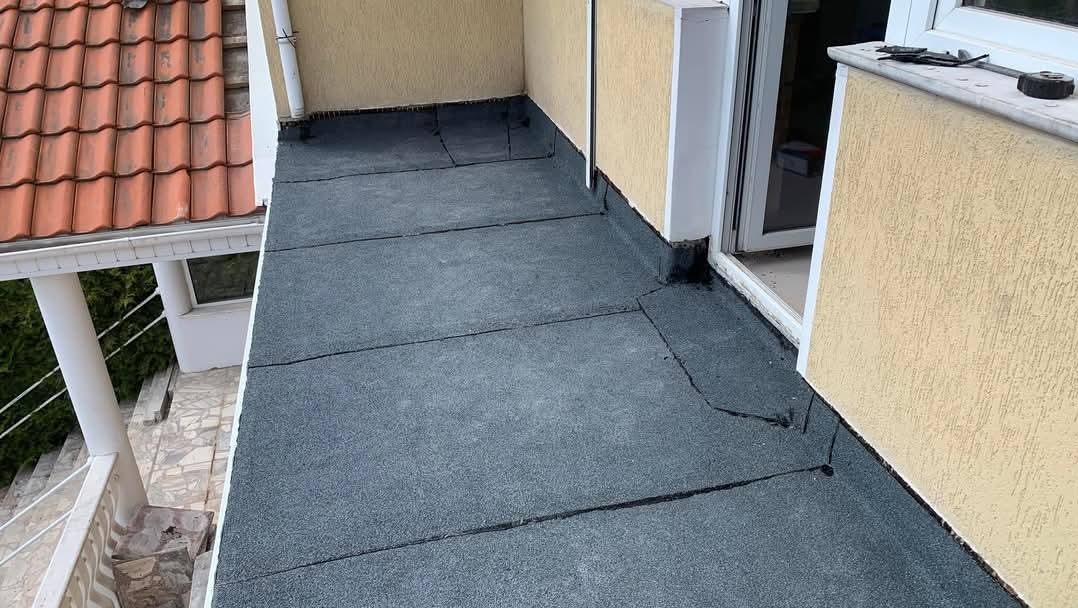 Wykończony balkon z szarą, bitumiczną izolacją, widok z góry. Fragment dachu z czerwoną dachówką w tle. Narzędzia na parapecie.