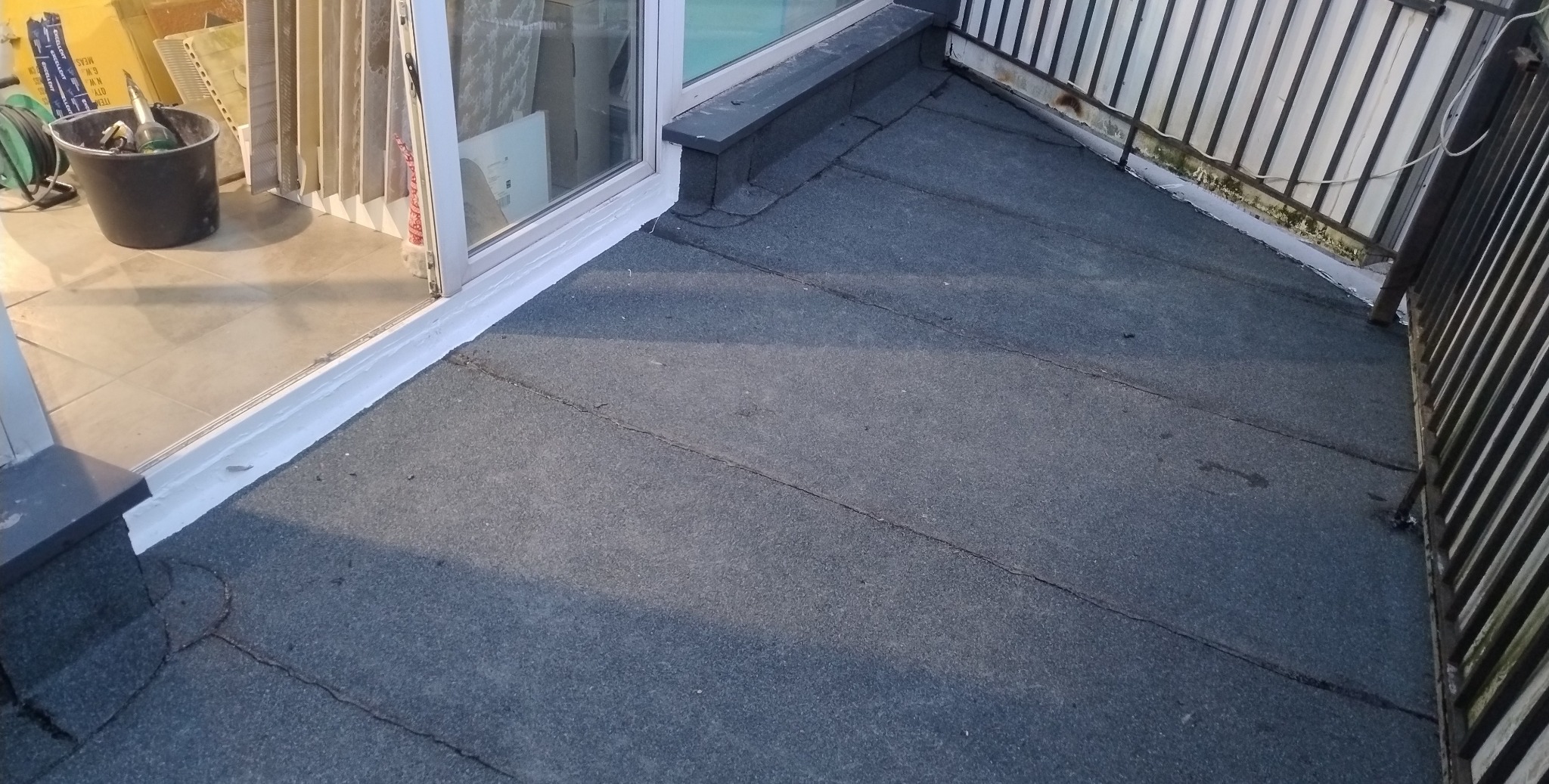 Wykończony balkon z bitumiczną izolacją, widoczne detale uszczelnienia przy oknie i balustradzie. Wiadro z narzędziami w rogu. Szare wykończenie, prosta forma.