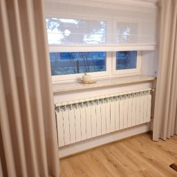 KATARZYNA CZADO - Biała roleta rzymska w oknie z beżowymi zasłonami po bokach, kremowy parapet z dekoracją i biały grzejnik. Widok na zewnątrz przez okno.