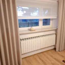 Biała roleta rzymska w oknie z beżowymi zasłonami po bokach, kremowy parapet z dekoracją i biały grzejnik. Widok na zewnątrz przez okno.