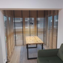 KATARZYNA CZADO - Wnętrze z drewnianymi żaluzjami poziomymi w oknach, stolik z blatem z litego drewna i fragment sofy w odcieniach zieleni.