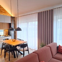 KATARZYNA CZADO - Nowoczesne zasłony w odcieniach beżu i bieli, upiększające przestrzeń jadalni z drewnianym stołem i czarnymi krzesłami. Ciepłe światło podkreśla ceglaną ścianę w tle.