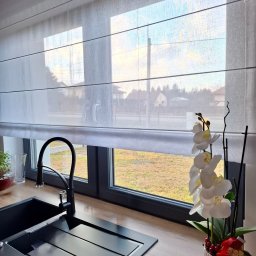 KATARZYNA CZADO - Jasna, zwiewna roleta rzymska w kuchni z czarnym zlewem i kwiatami. Widok z okna na zewnątrz. Delikatne światło dzienne.