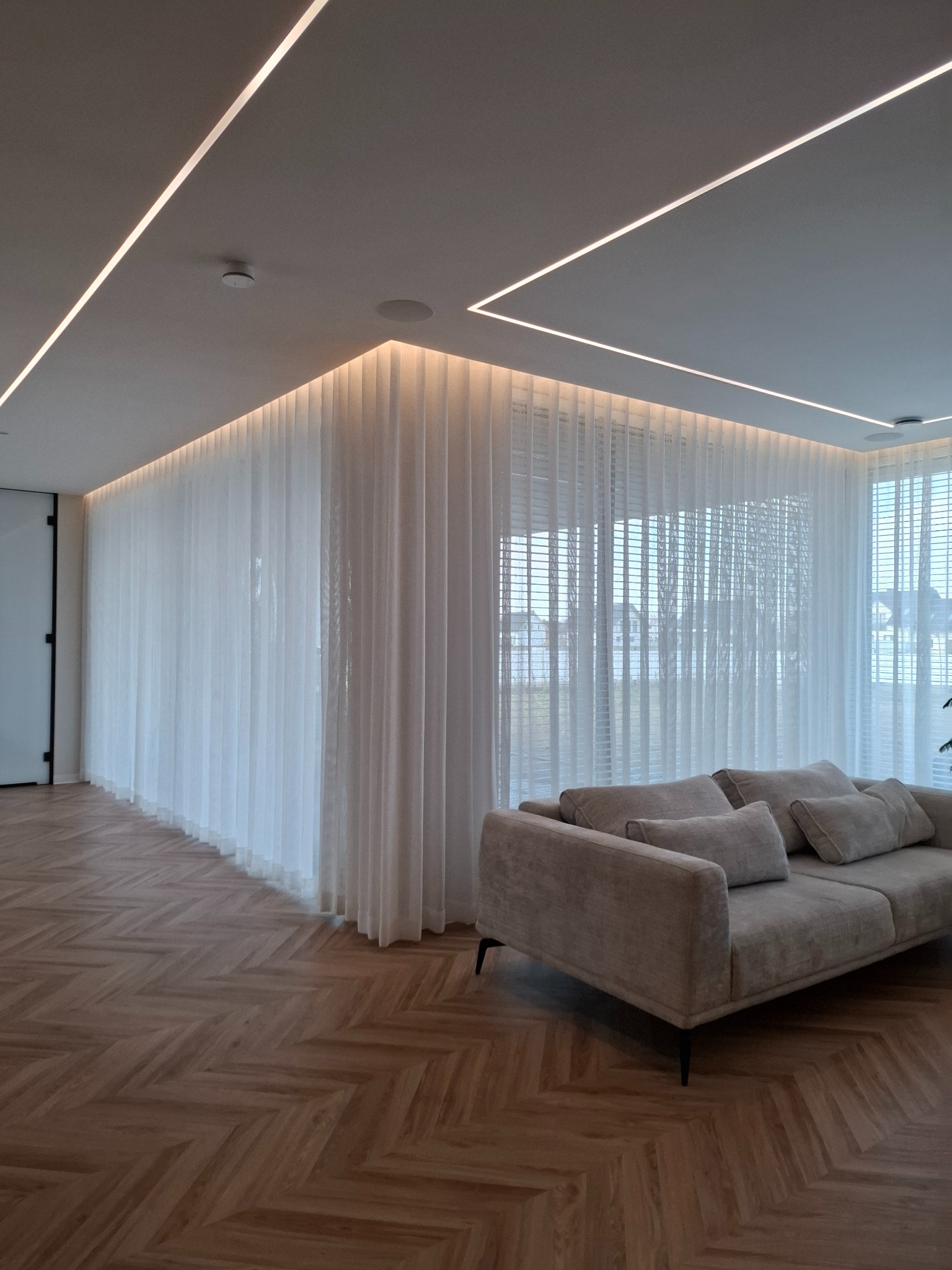 Eleganckie, długie, białe zasłony w nowoczesnym salonie z parkietem w jodełkę i podświetlanym sufitem. Widoczne także żaluzje w oknach.