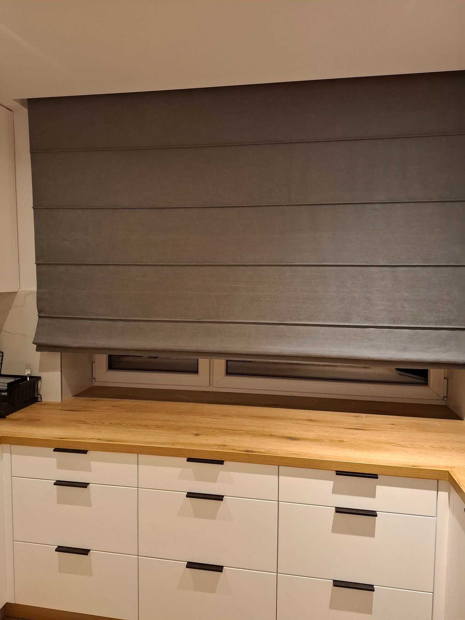 Szara roleta rzymska zamontowana w kuchni, widoczna nad blatem i oknem. Minimalistyczny design, jasne meble kuchenne z ciemnymi uchwytami.