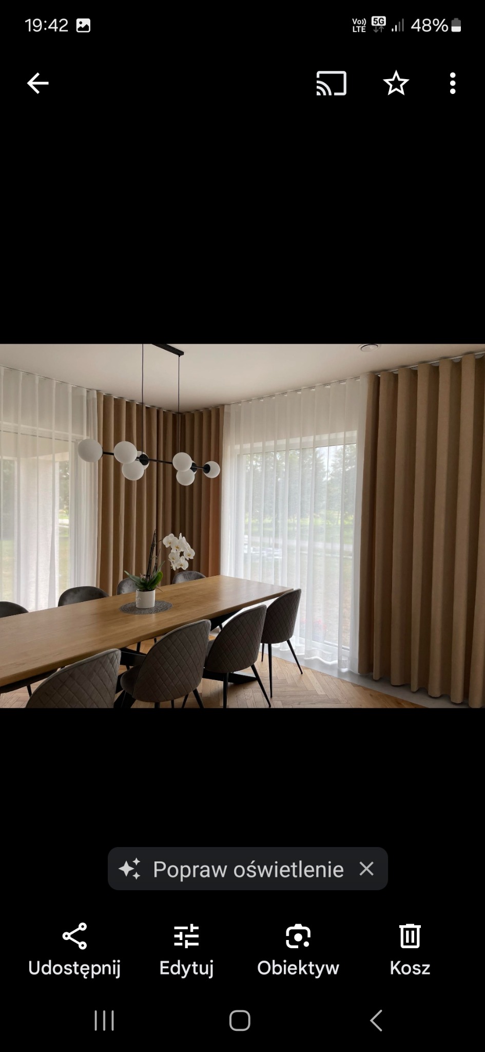 Elegancka jadalnia z drewnianym stołem i krzesłami, oświetlona lampą wiszącą. Okna zdobią beżowe, grube zasłony oraz delikatne, białe firany.