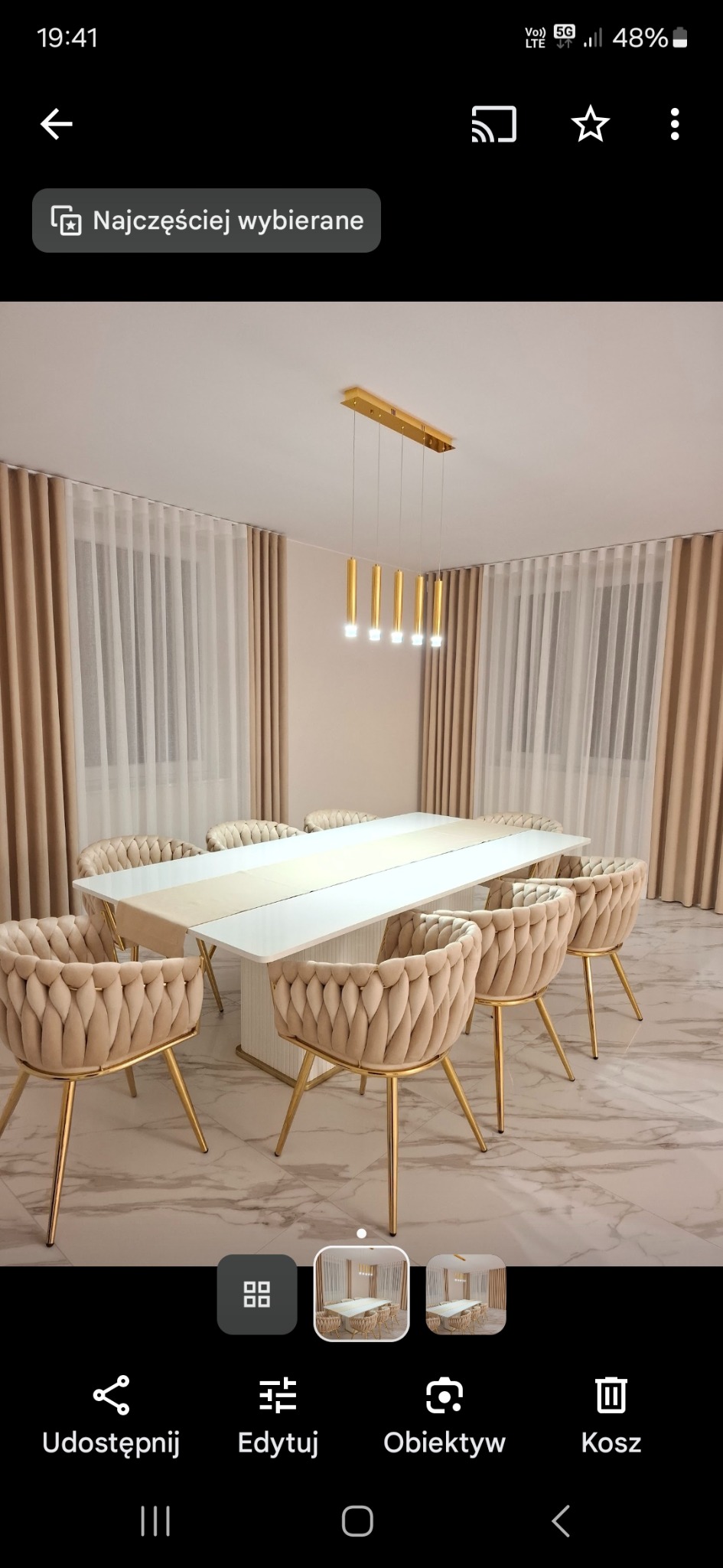 Beżowe zasłony w jadalni z białym stołem i krzesłami na złotych nogach. Złota lampa wisząca nad stołem. Marmurowa podłoga.