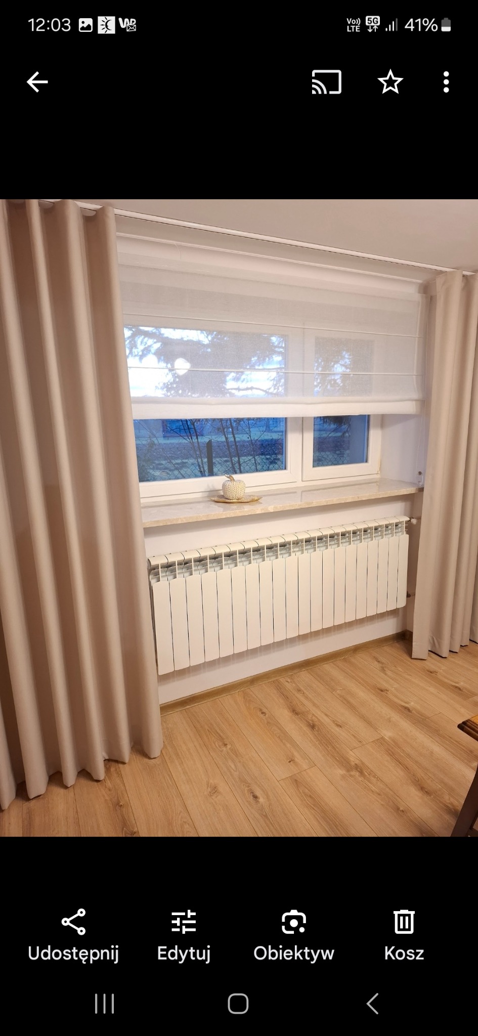 Biała roleta rzymska w oknie z beżowymi zasłonami po bokach, kremowy parapet z dekoracją i biały grzejnik. Widok na zewnątrz przez okno.