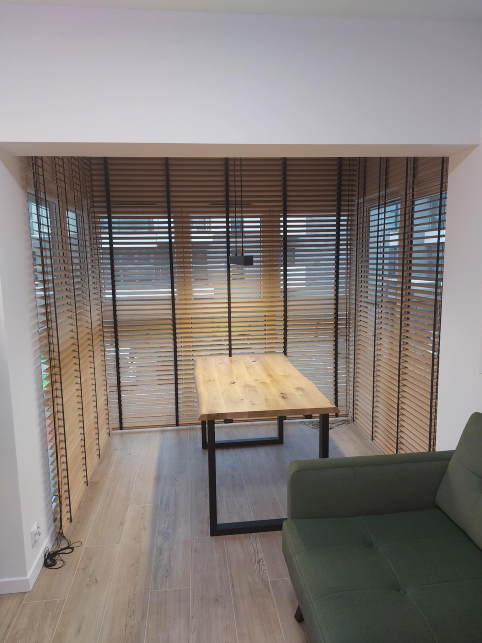 Wnętrze z drewnianymi żaluzjami poziomymi w oknach, stolik z blatem z litego drewna i fragment sofy w odcieniach zieleni.