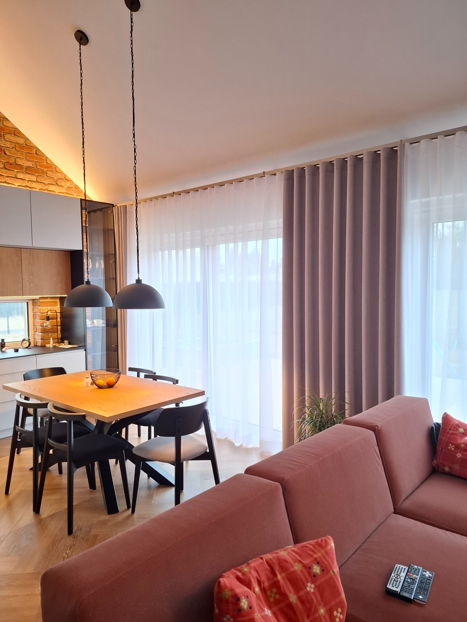 Nowoczesne zasłony w odcieniach beżu i bieli, upiększające przestrzeń jadalni z drewnianym stołem i czarnymi krzesłami. Ciepłe światło podkreśla ceglaną ścianę w tle.